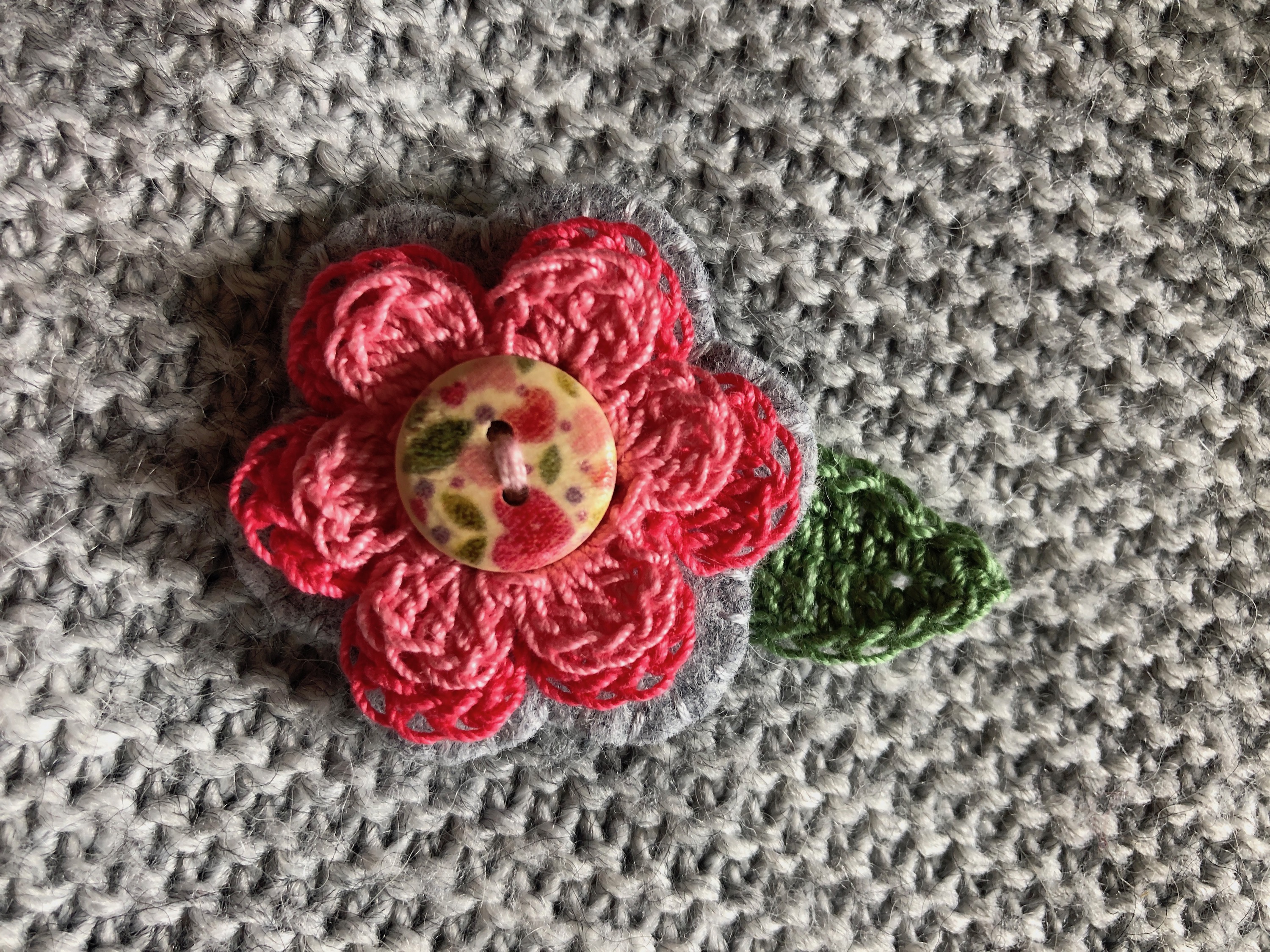 A hand stitched felt and crocheted flower brooch in pinks and pale grey with patterned button in centre.