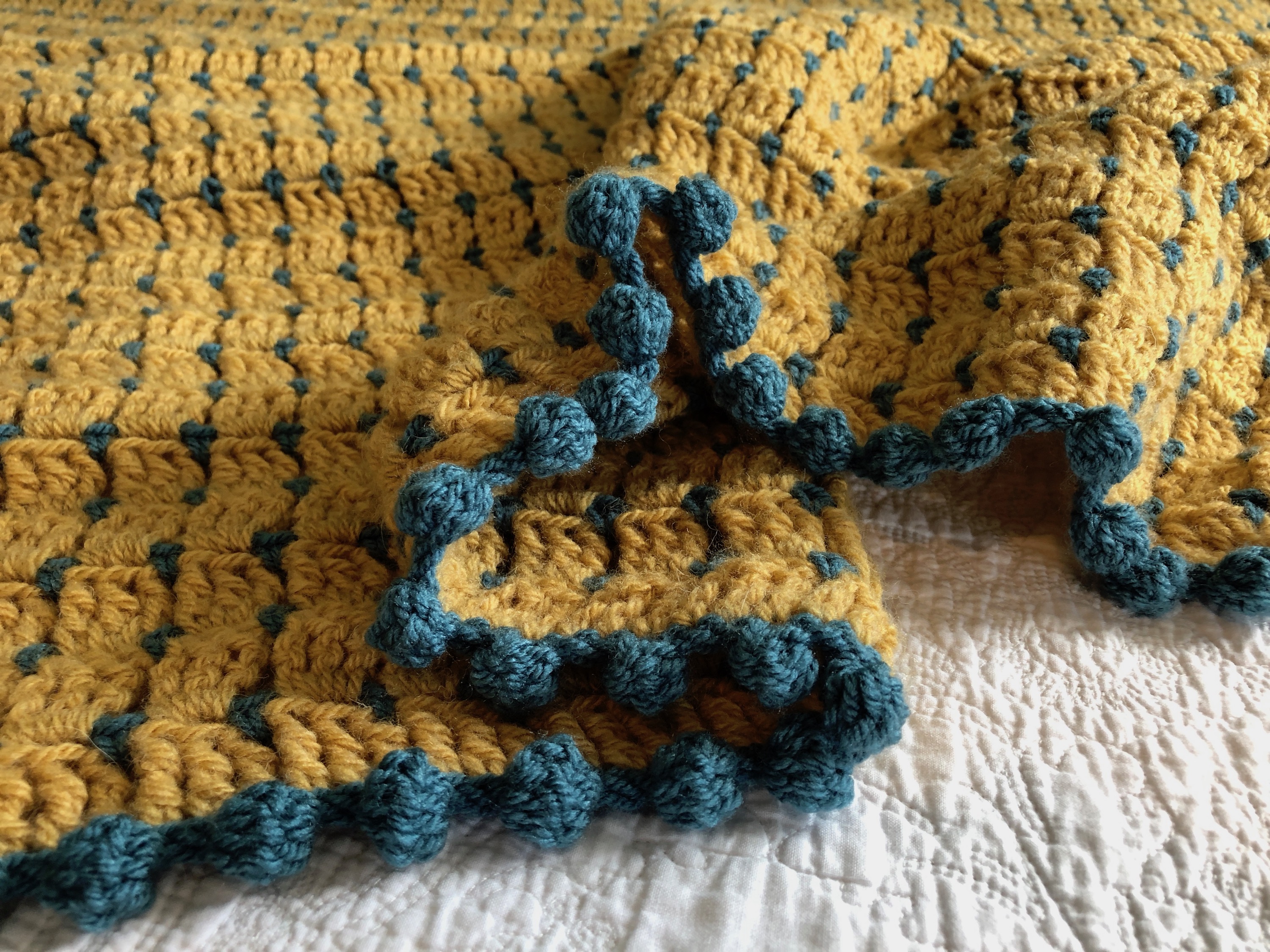 Block stitch blanket in mustard yellow and teal, with pom-pom edging.