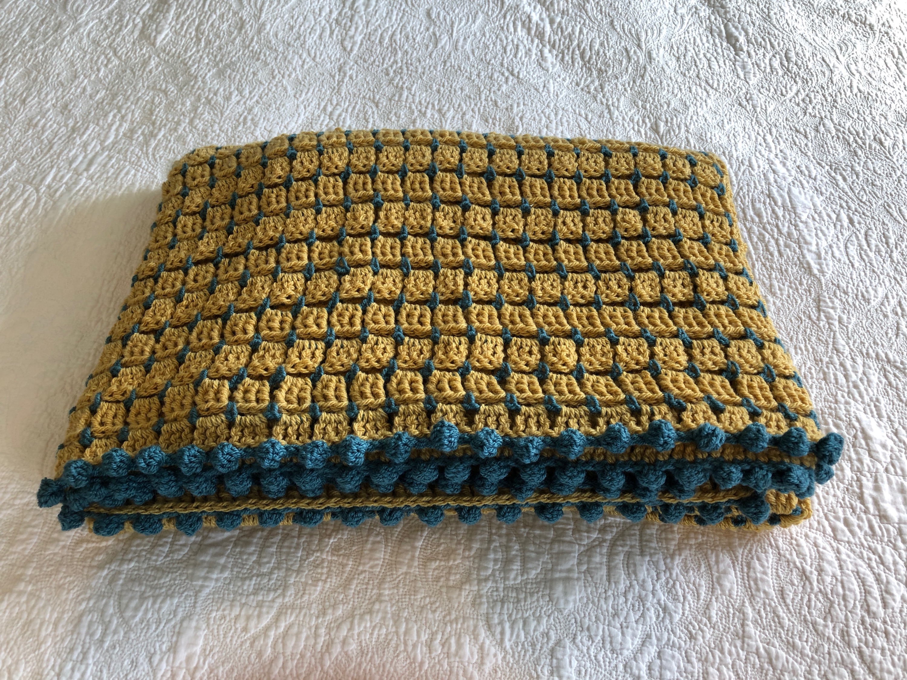 Block stitch blanket in mustard yellow and teal, with pom-pom edging.