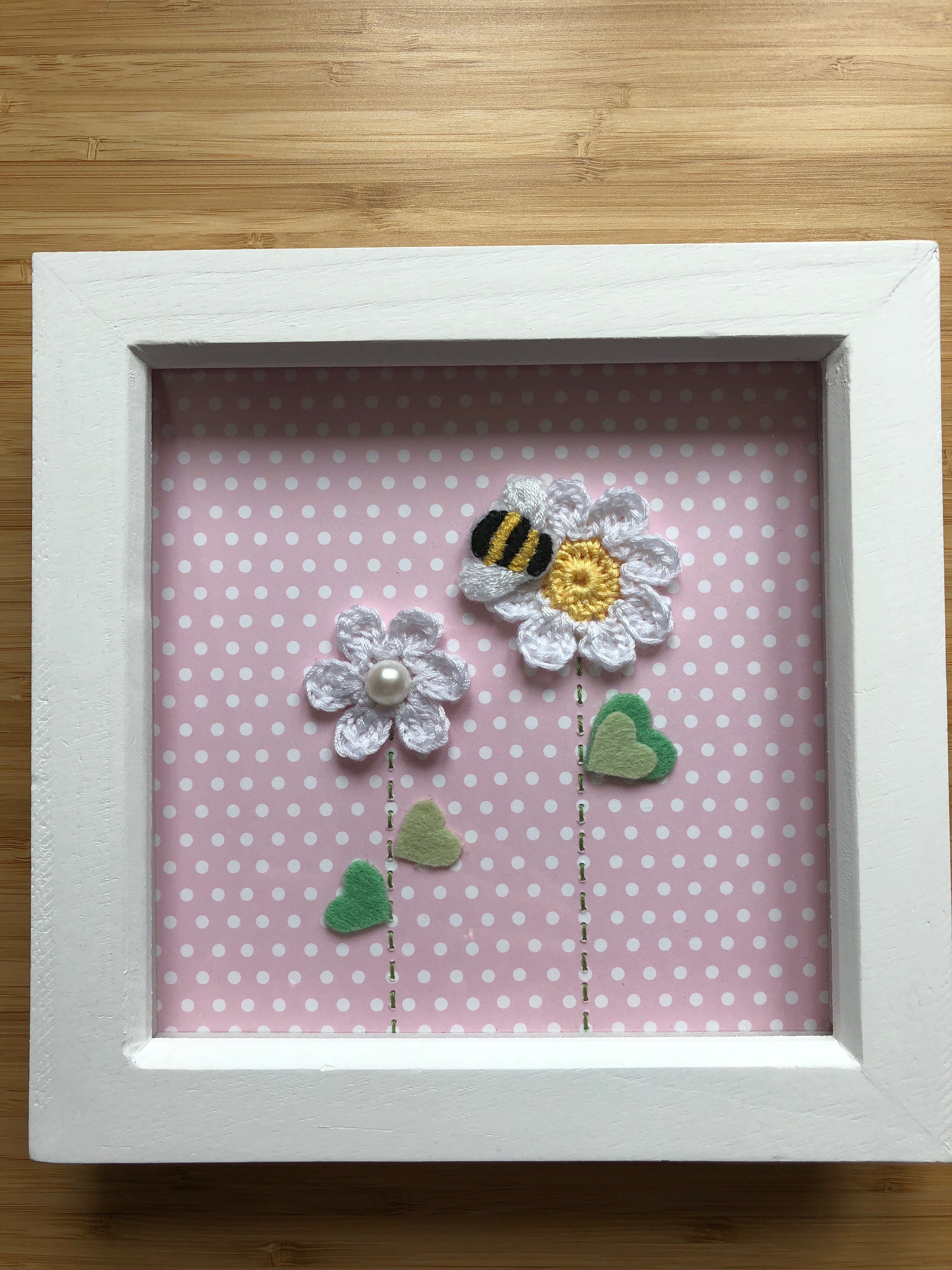 Hand crocheted white daisy flowers and hand embroidered bumblebee picture on pink polkadot background in a white wooden frame.