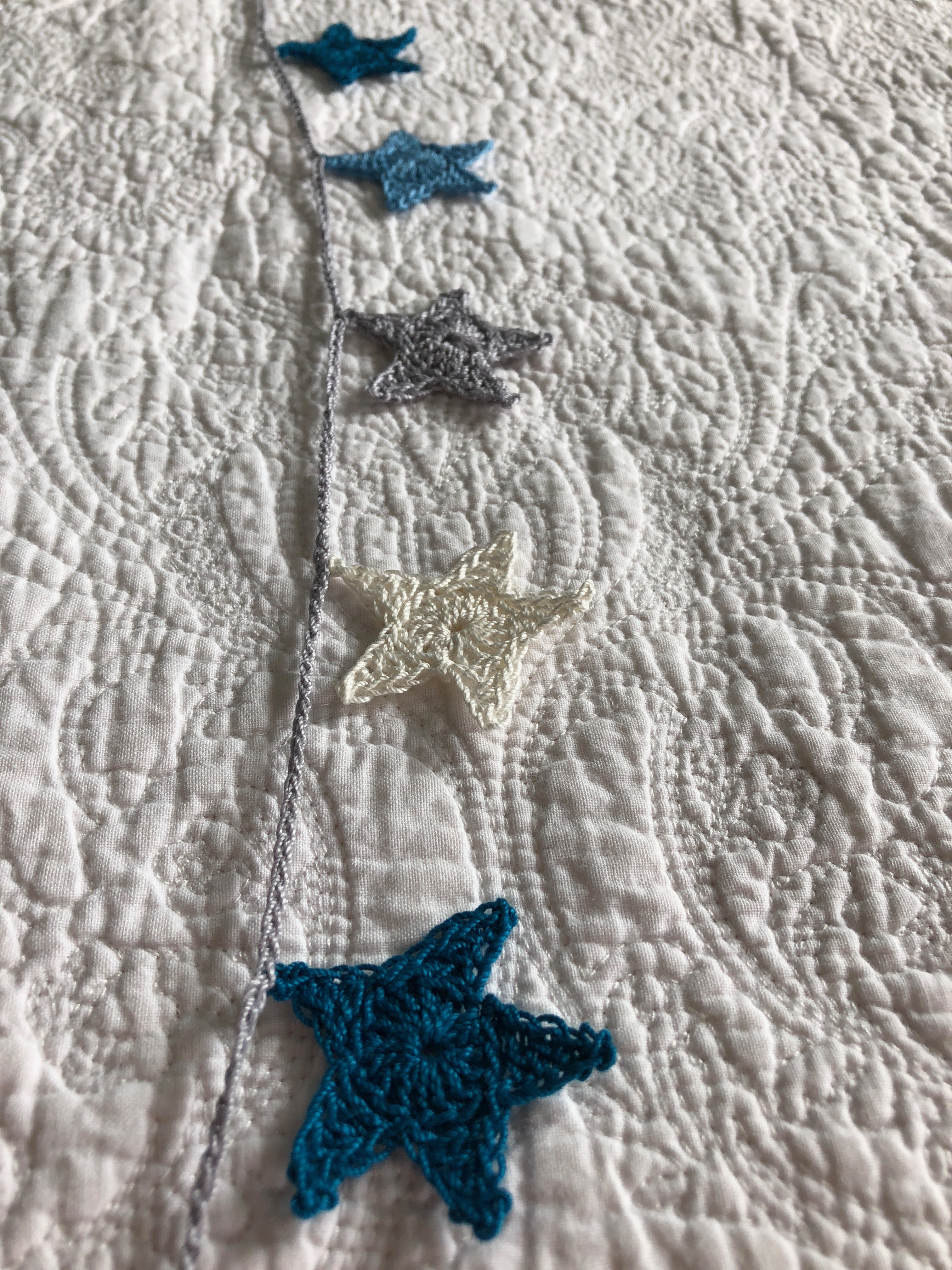 A small hand crocheted cotton star garland in petrol blue, mid blue, grey and white with a grey crocheted cord.