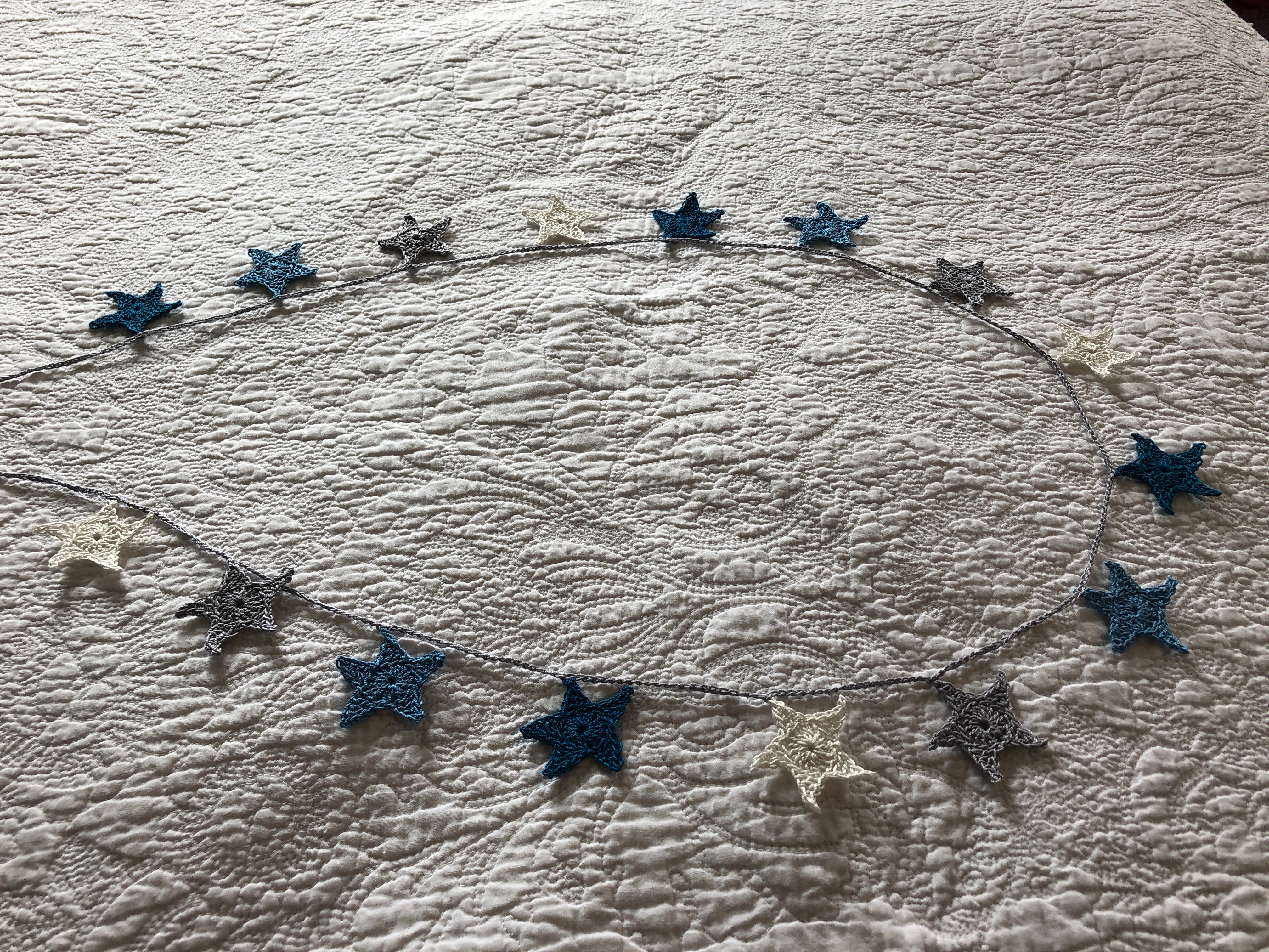 A small hand crocheted cotton star garland in petrol blue, mid blue, grey and white with a grey crocheted cord.