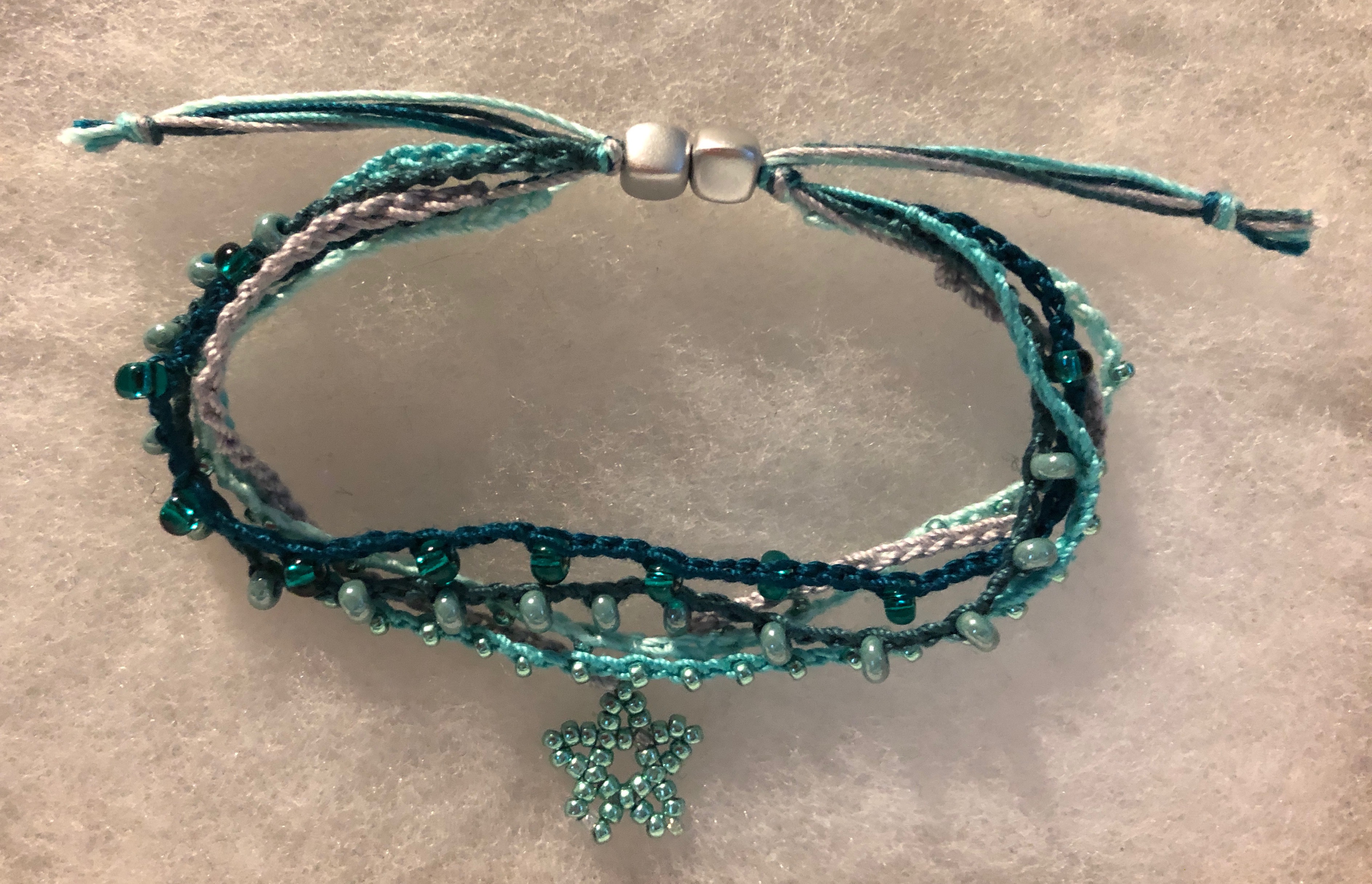 A handmade, crocheted, adjustable bracelet made with 5 strands of crocheted cotton and glass bead with and central beaded star hanging decoration. In a gradient of green shades.