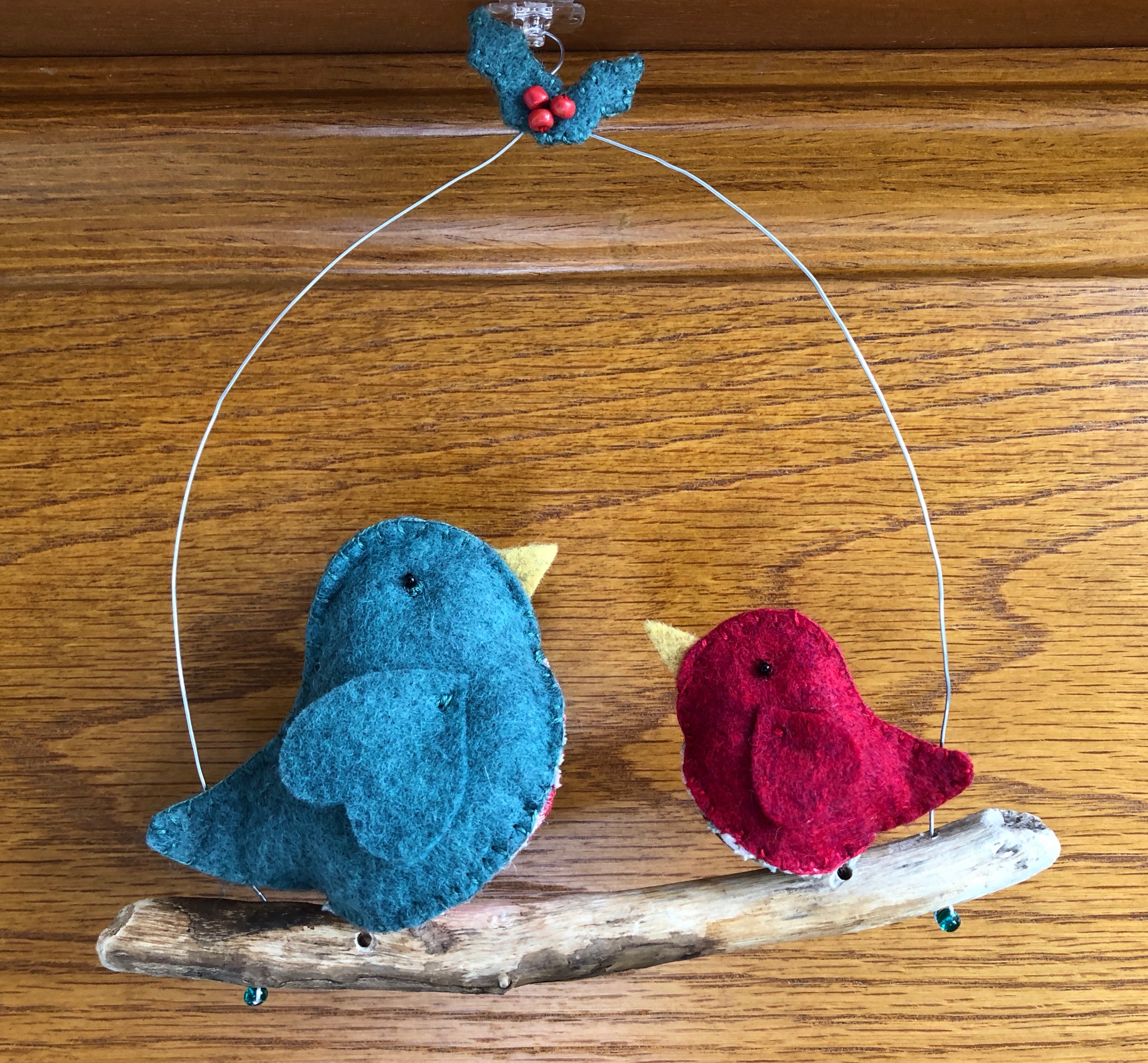 A medium and small pair of handmade, hand sewn, felt and fabric birds sat on a driftwood and wire hanging perch, with a tiny felt and bead holly decoration.