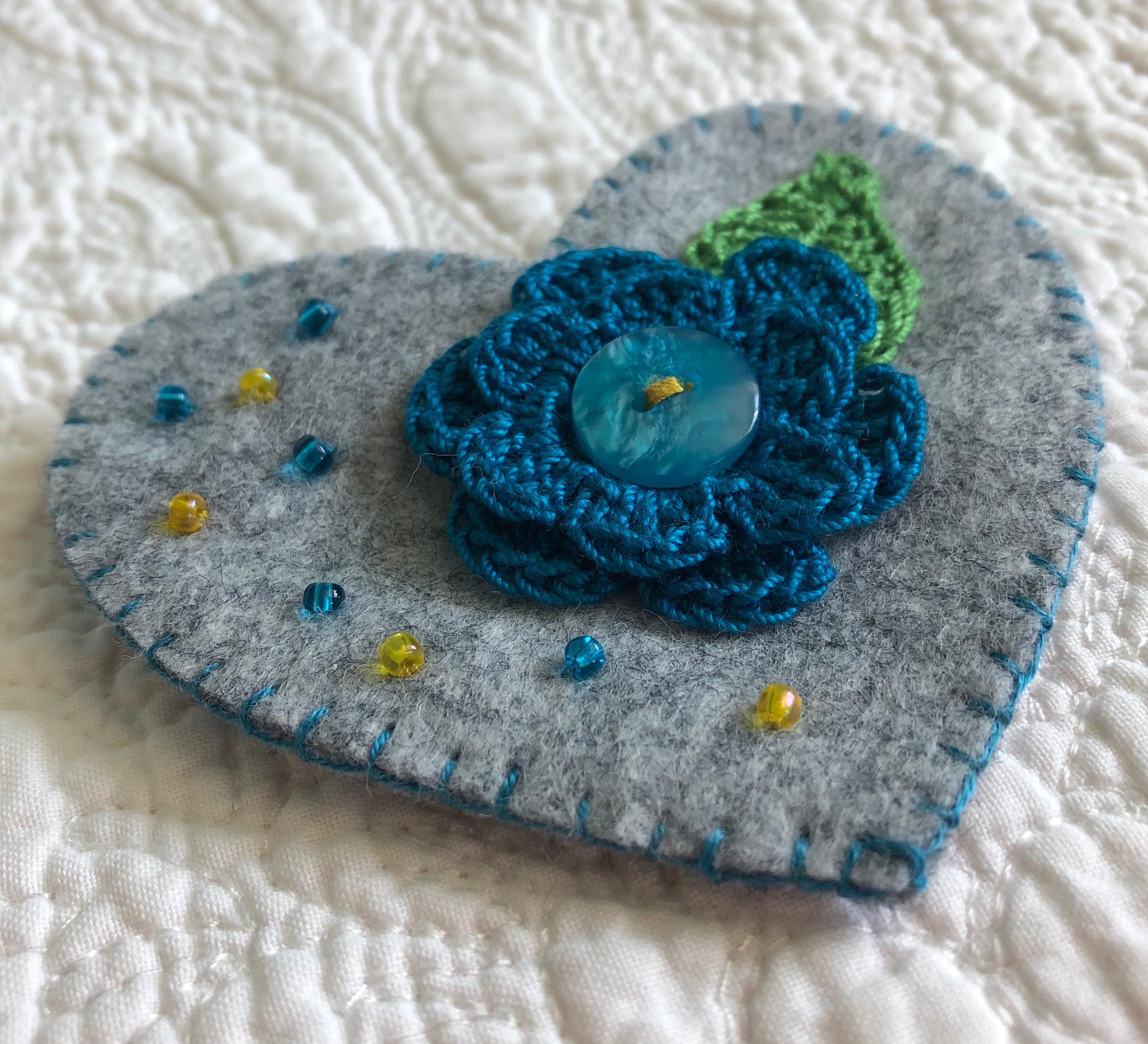 Handmade and hand stitched grey felt heart with a blue crocheted flower and green leaf embellished with a button and tiny glass beads.