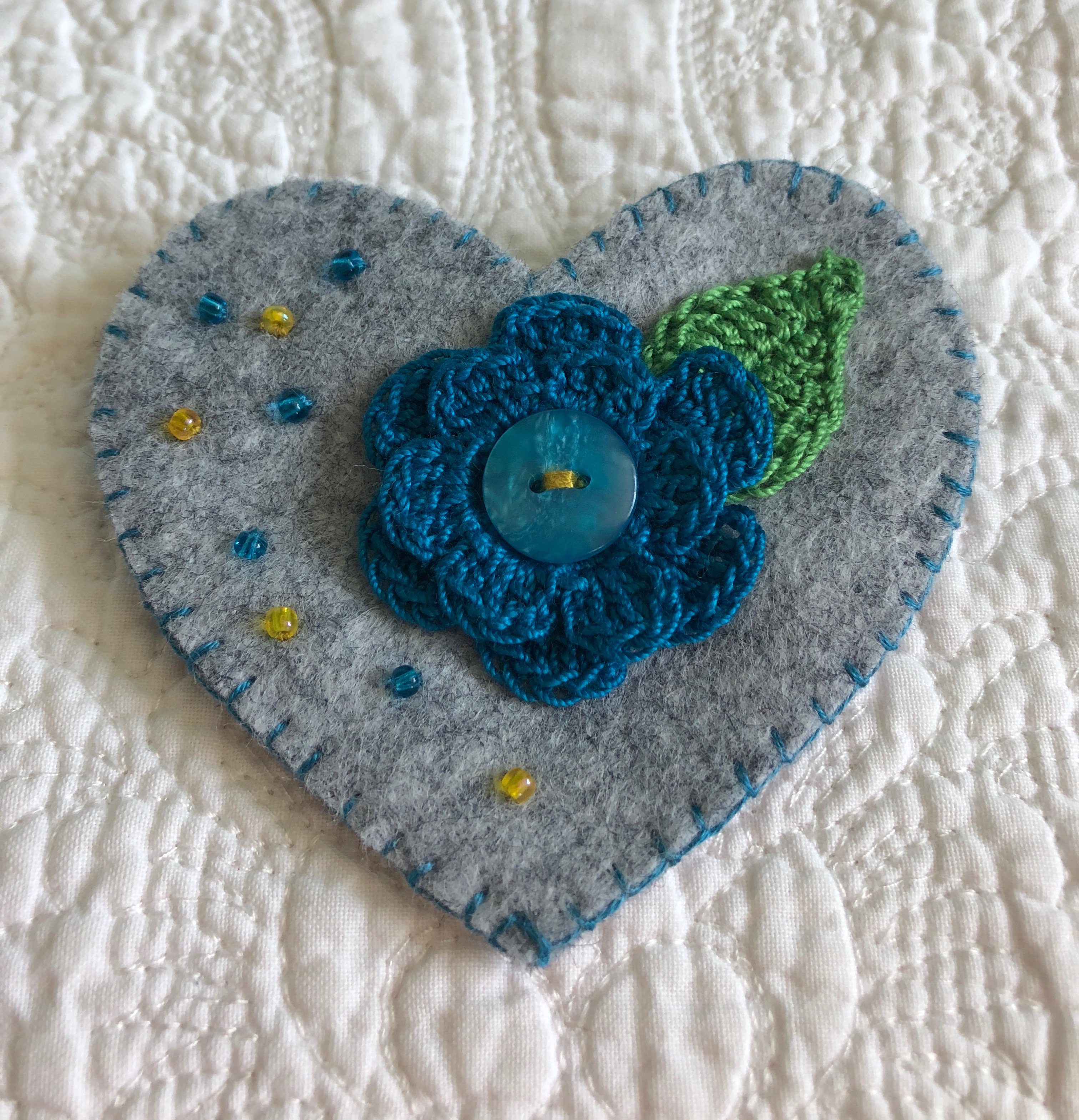 Handmade and hand stitched grey felt heart with a blue crocheted flower and green leaf embellished with a button and tiny glass beads.