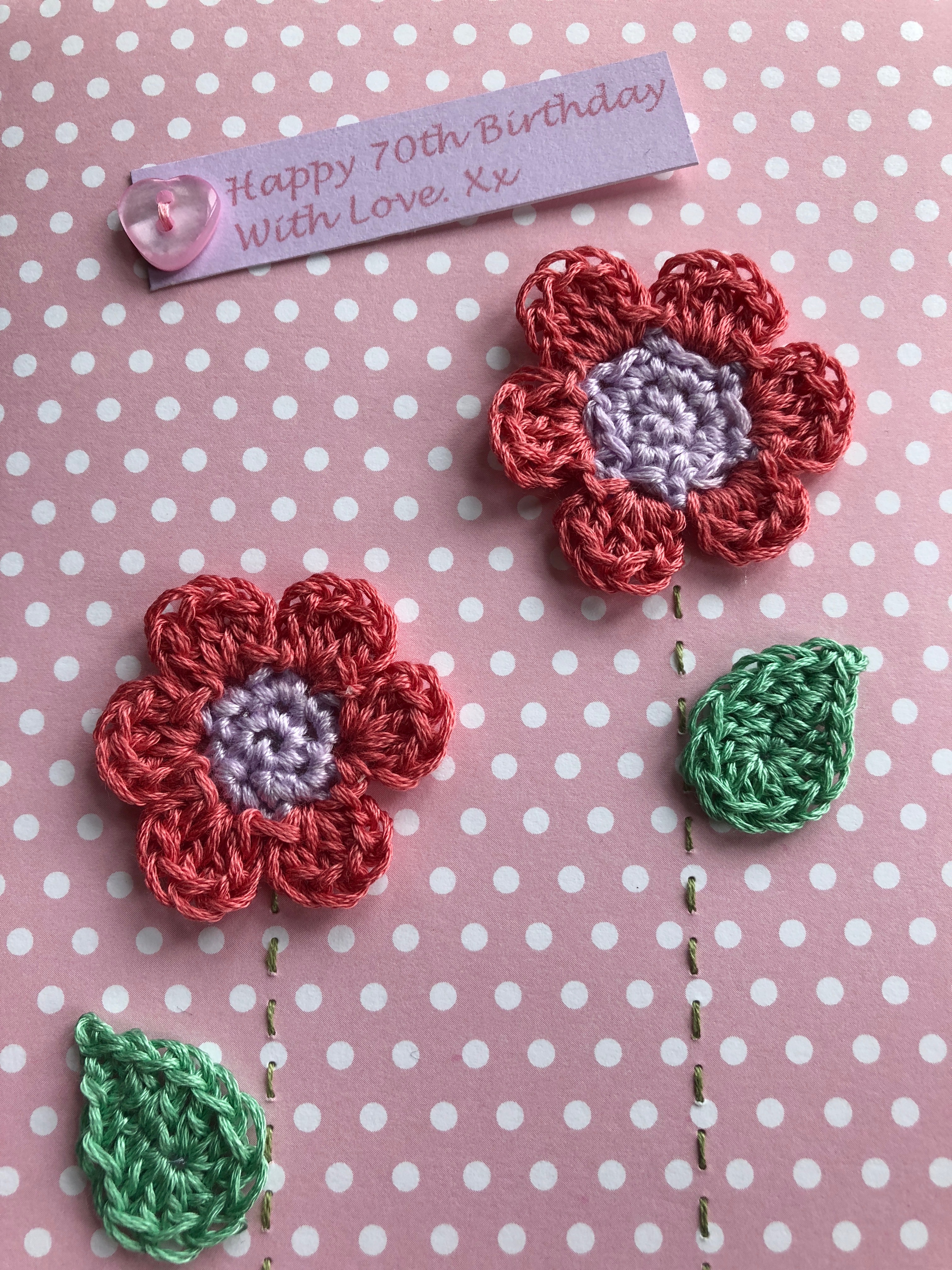 A handmade, crocheted and stitched card with two pink and lilac crocheted flowers with green crocheted leaves and a printed message with hand sewn button detail.