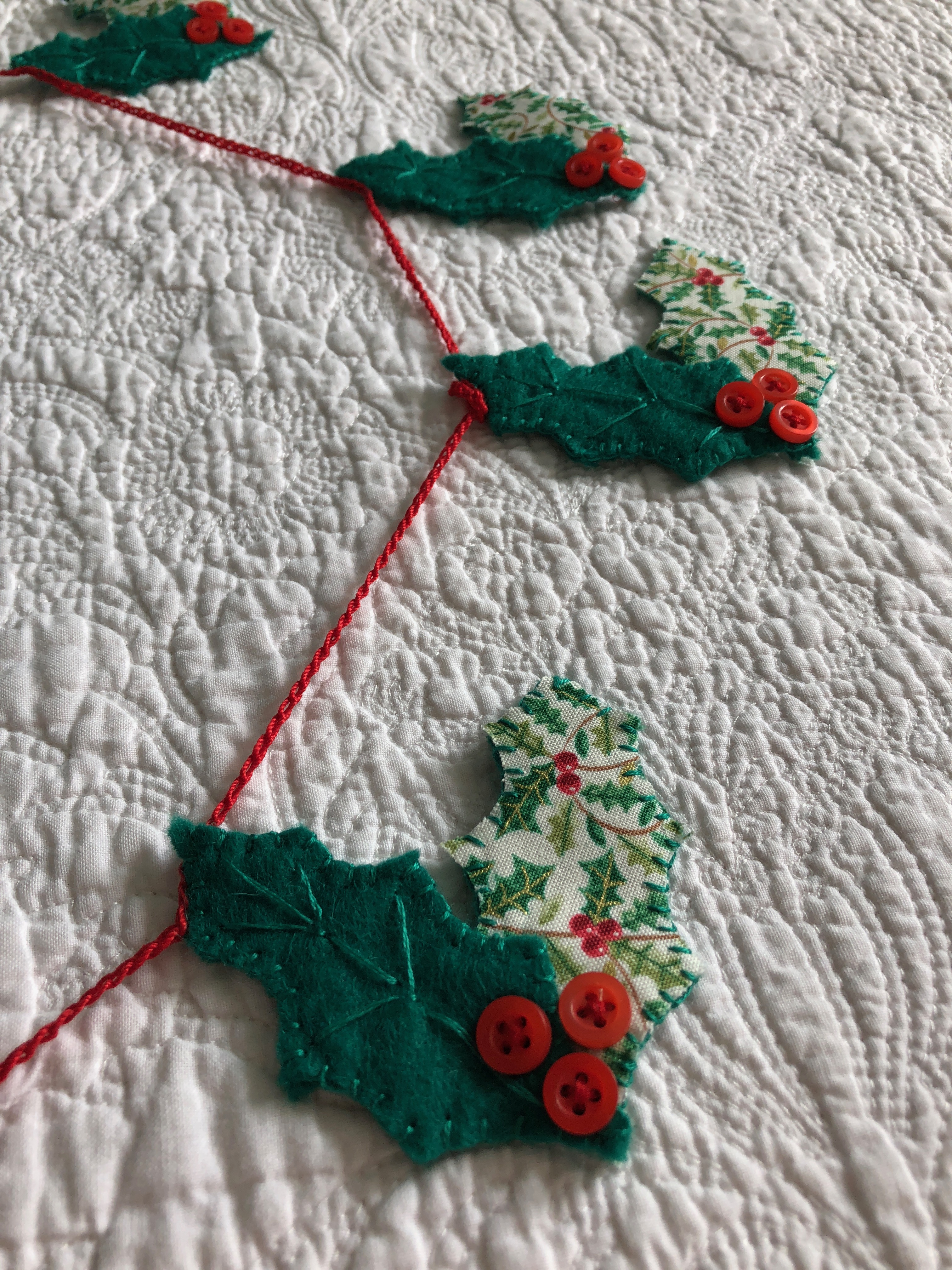 A handmade, hand stitched garland of green felt and holly printed cotton fabric holly leaves with red button berries.