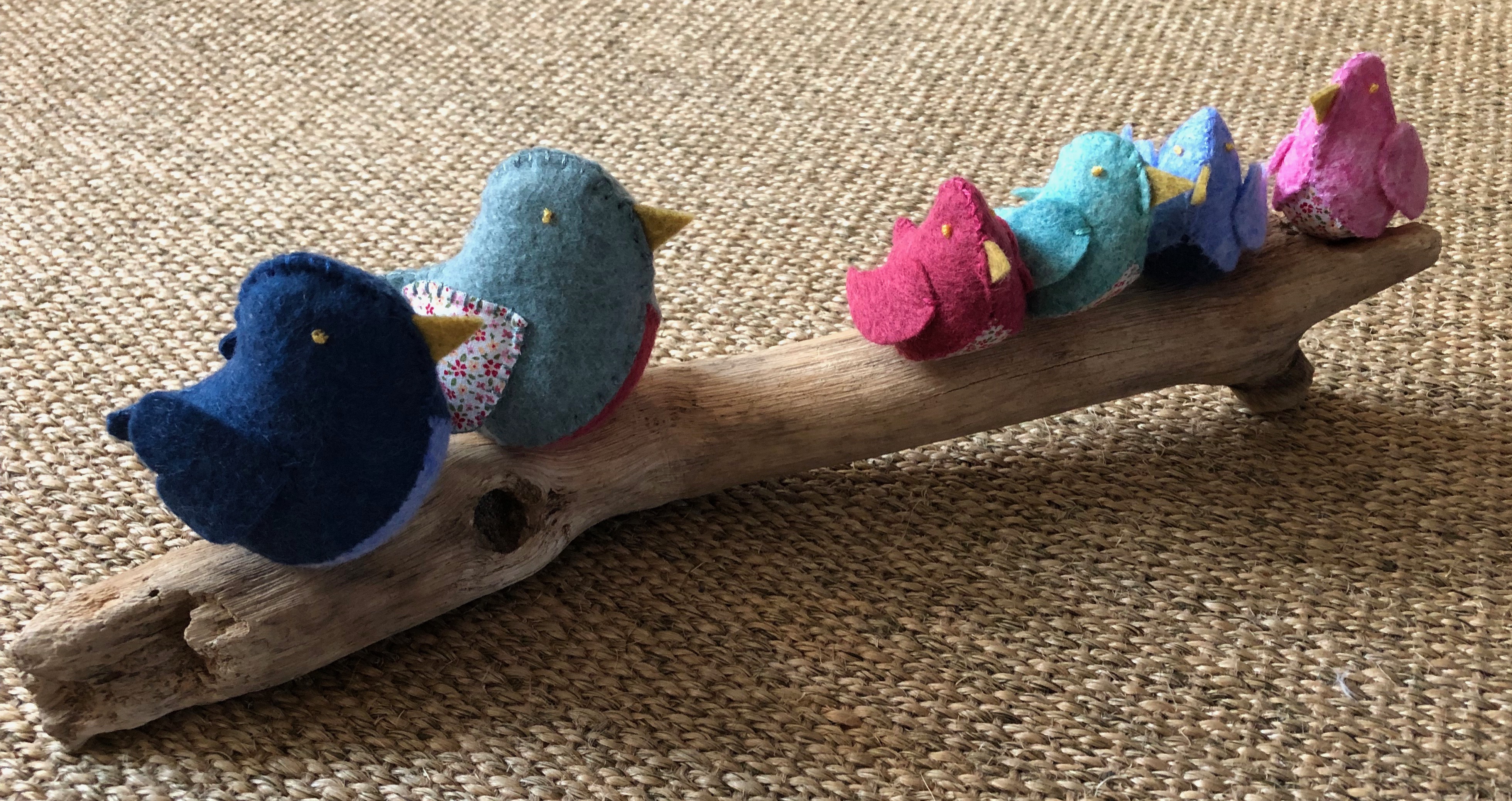 A family of 6 handmade and hand stitched felt and fabric birds sat on a drift wood log. Birds are made in a range of complimentary pastel shades of blue green and pink with tiny flower fabric chests or wings.