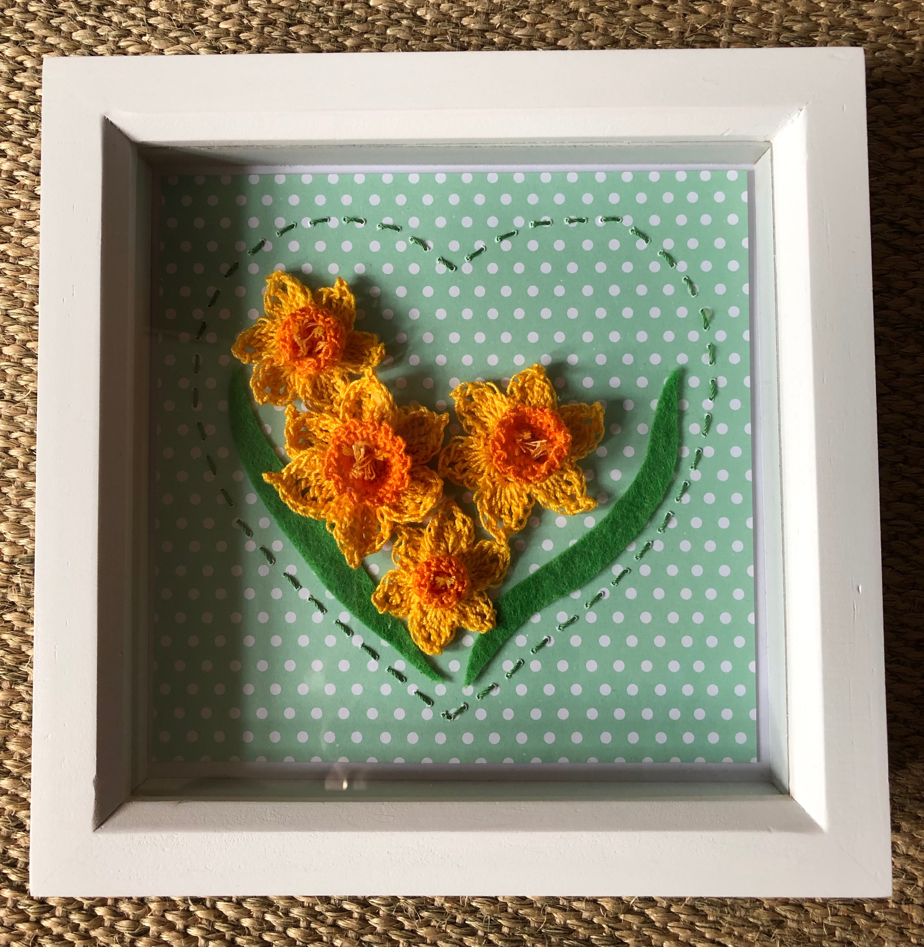 A white wooden framed picture with 4 handmade, crocheted yellow and orange daffodils with green felt leaves surrounded by a hand stitched green heart on a green and white spotty background.