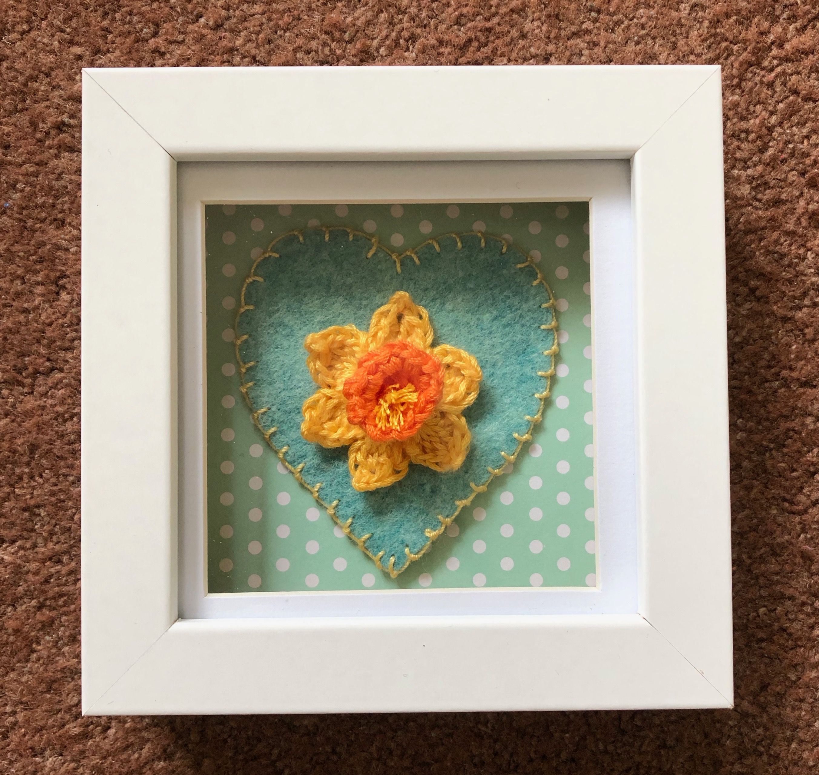 A small white wooden framed picture with a single handmade, crocheted yellow and orange daffodil on top of a hand stitched green and yellow felt heart on a green and white spotty background.