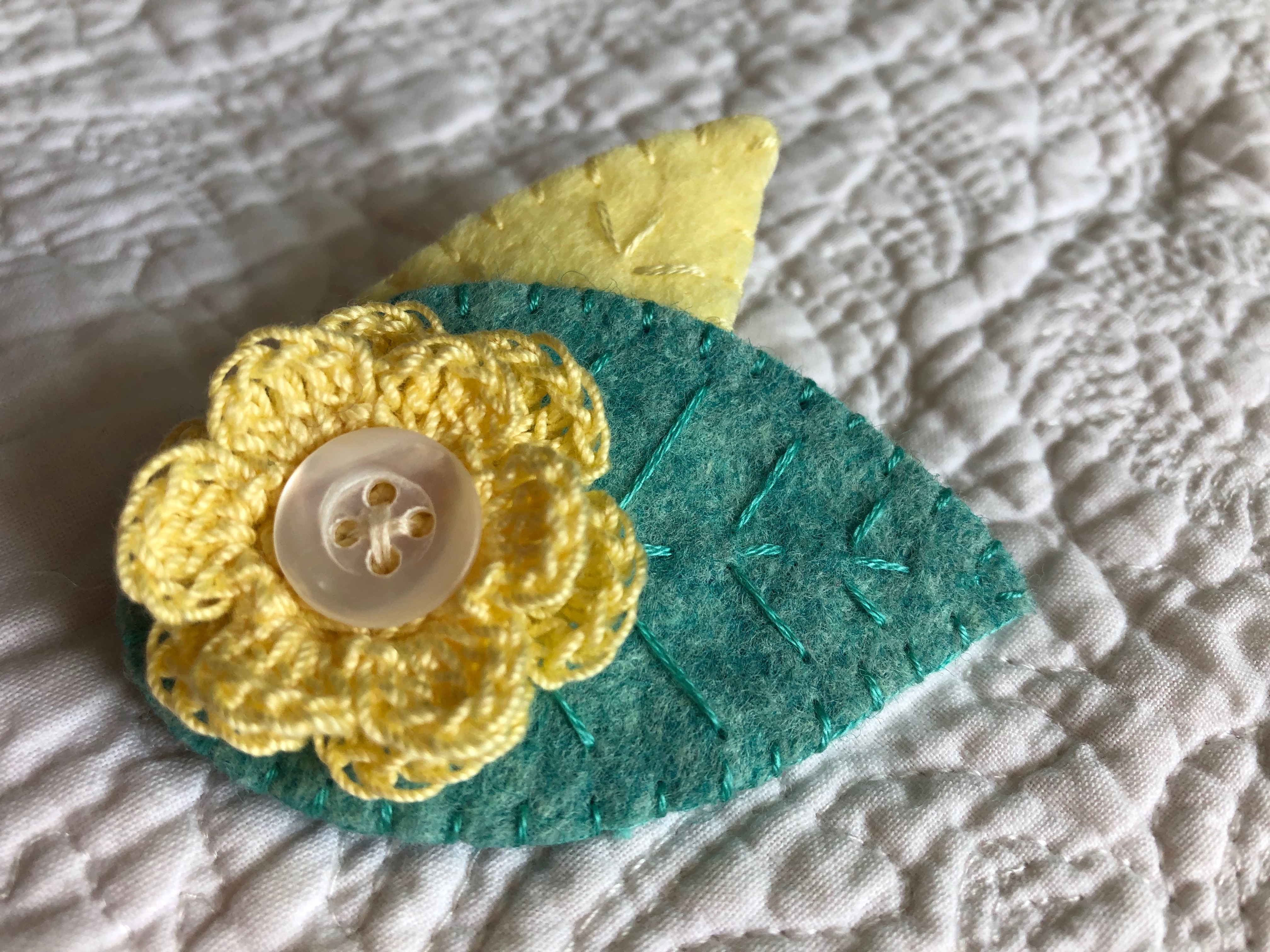 A handmade felt and crocheted brooch with hand stitched and button detailing. Pale yellow flower with pale yellow and green hand detailed felt leaves.