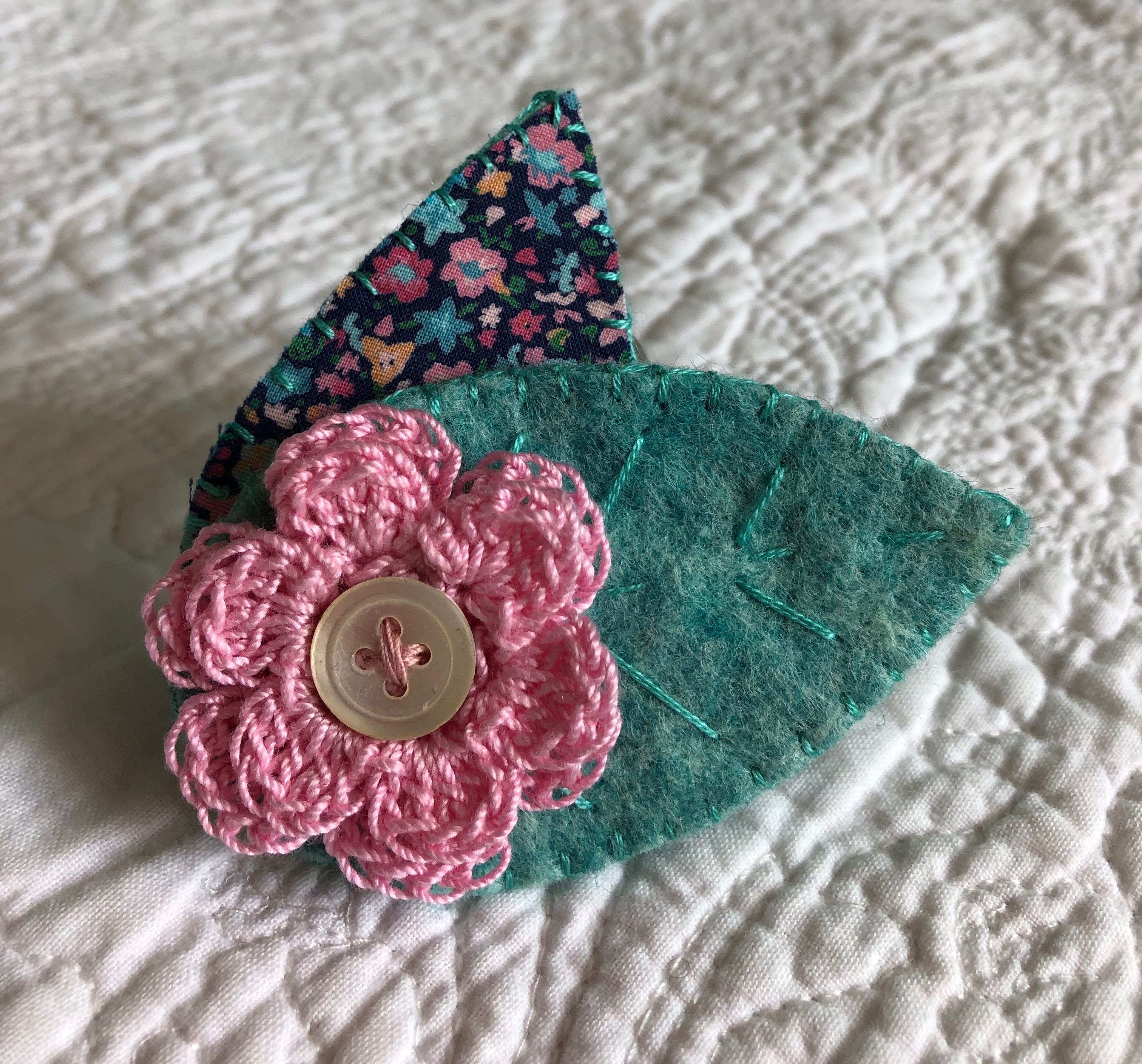 A handmade cotton fabric, felt and crocheted brooch with hand stitched and button detailing. Pink flower with multicoloured floral fabric leaf and hand detailed green felt leaf.