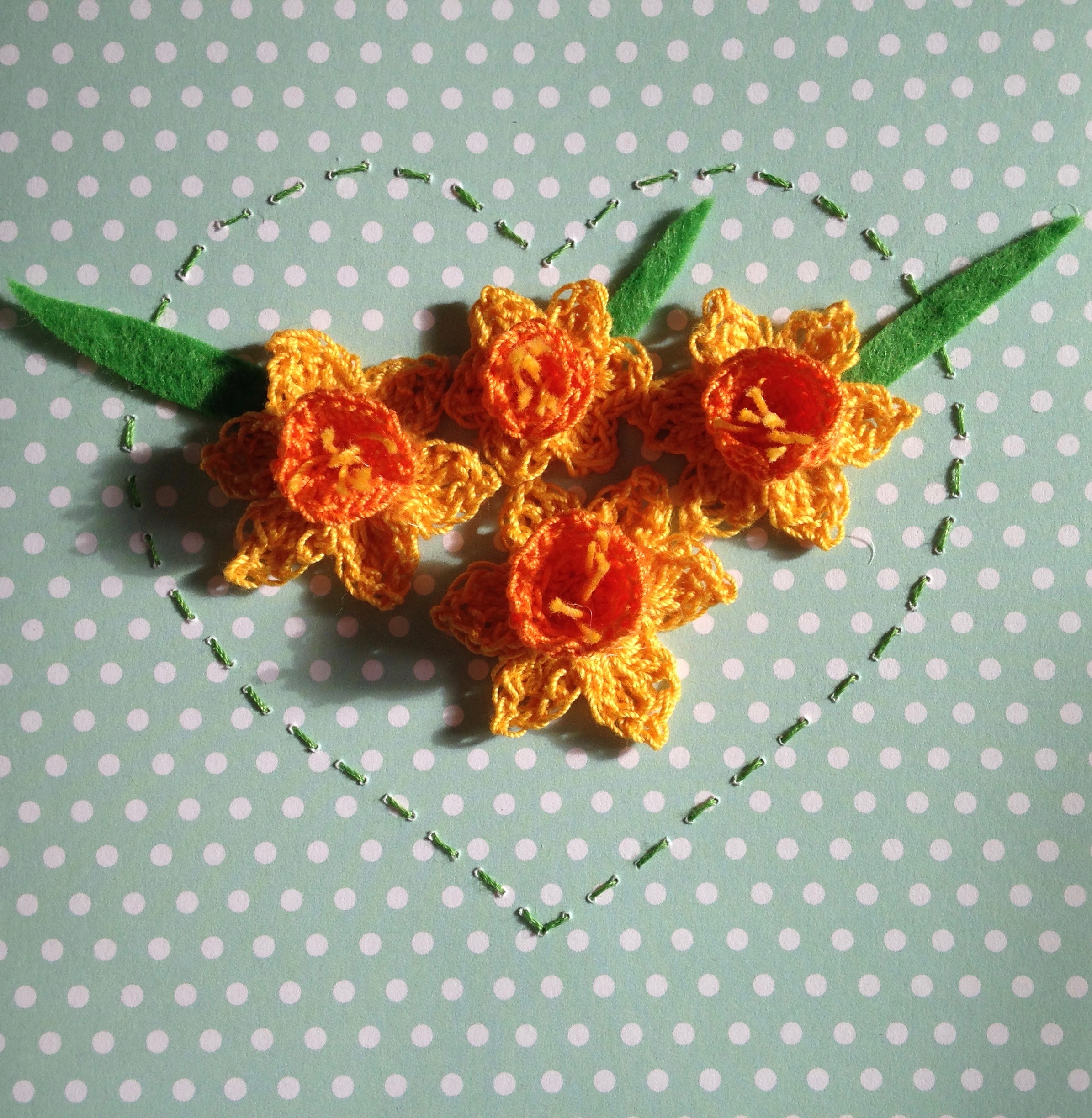 A crocheted Daffodil card. Crocheted Daffodils in yellow and orange surrounded by a hand stitched heart. With a green and white polka dot card background.