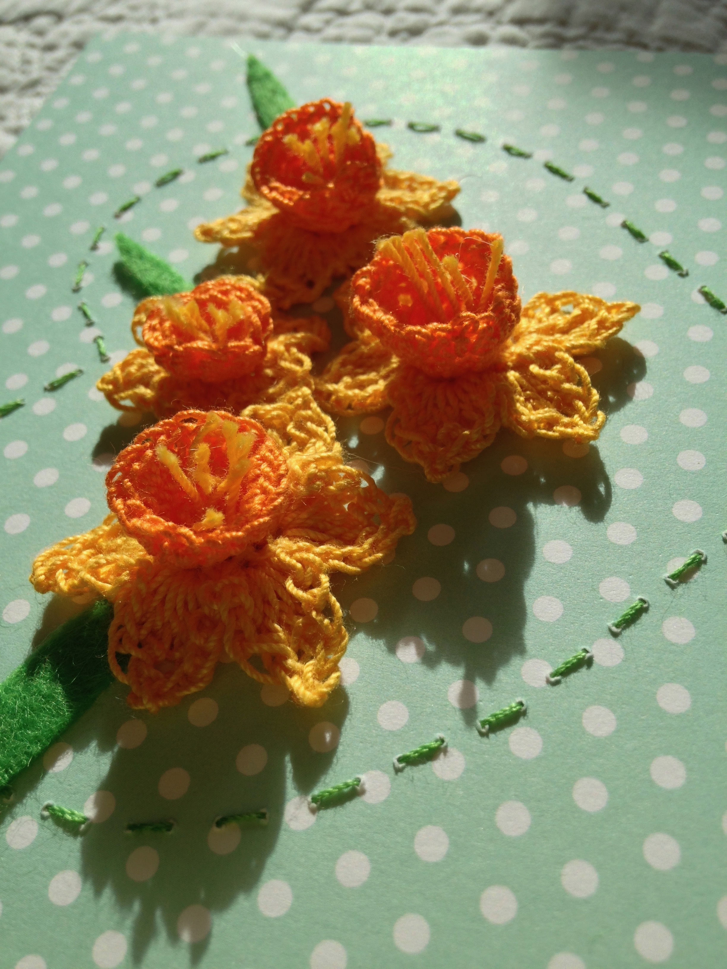 A crocheted Daffodil card. Crocheted Daffodils in yellow and orange surrounded by a hand stitched heart. With a green and white polka dot card background.