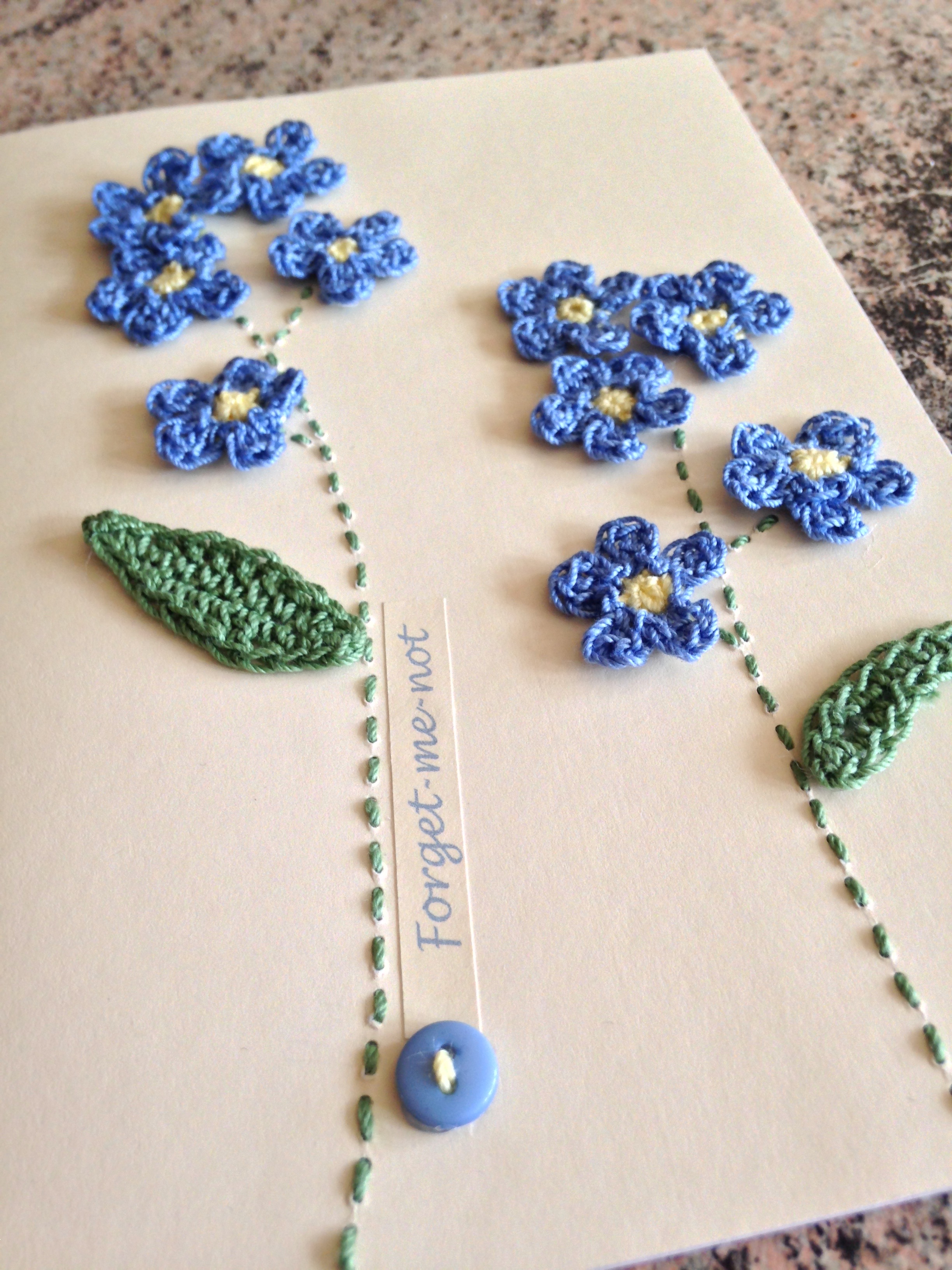 A hand stitched and crocheted greetings card with pale blue Forget-me-not flowers and green leaves.