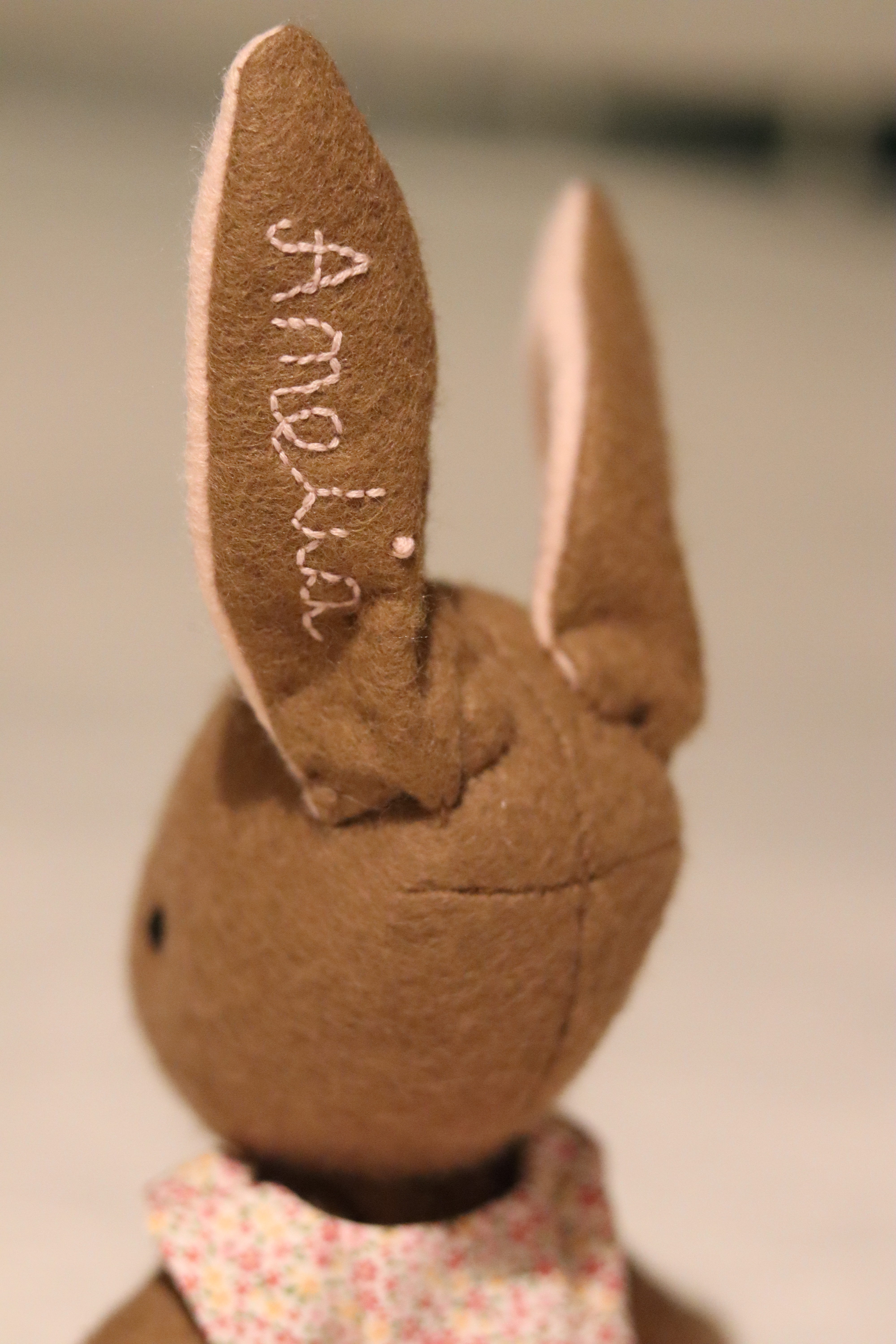 A handmade brown felt bunny rabbit with cotton floral dress and a natural linen coat with a hand embroidered name on her ear. Made using a pattern by Simone Gooding.