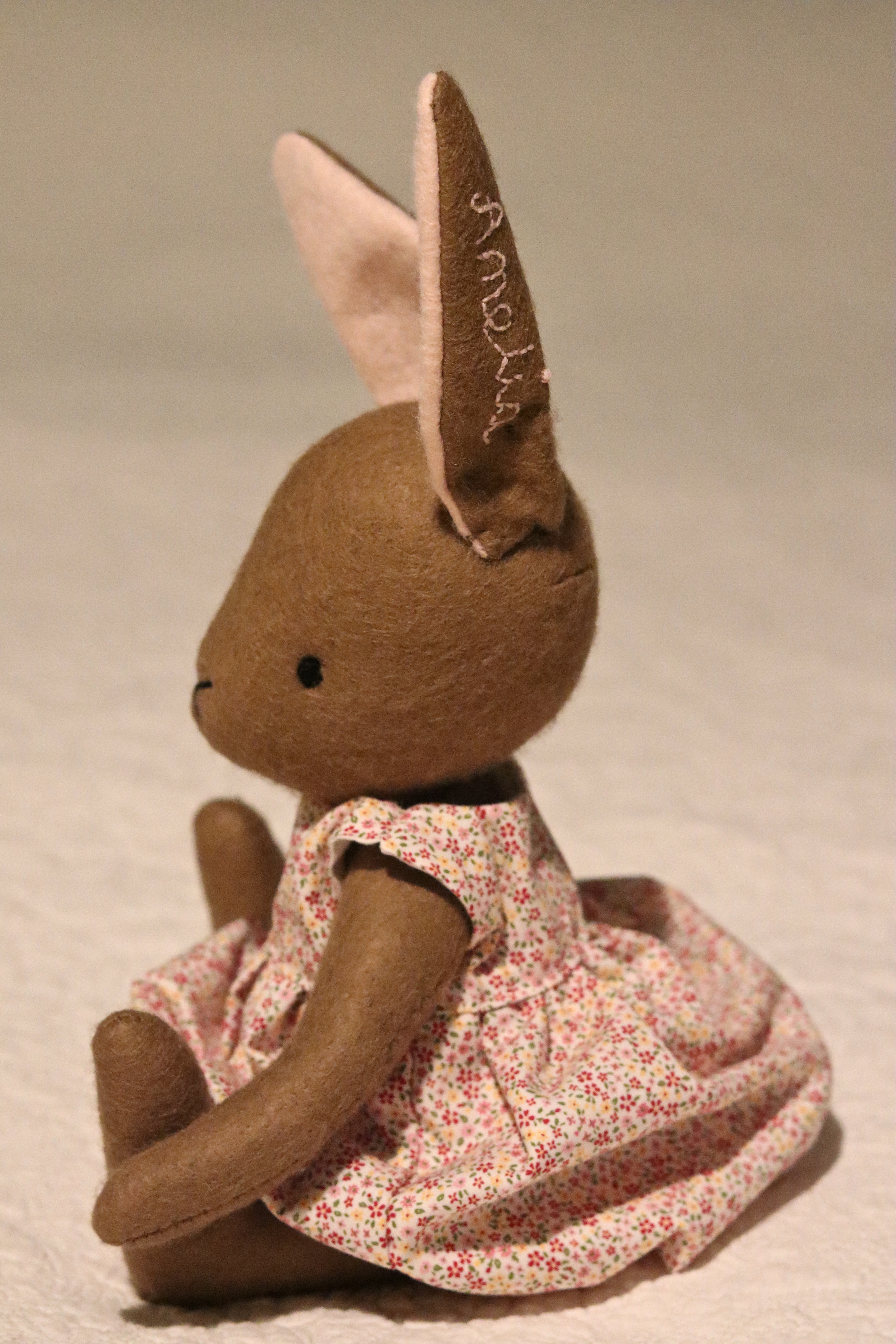 A handmade brown felt bunny rabbit with cotton floral dress and a natural linen coat with a hand embroidered name on her ear. Made using a pattern by Simone Gooding.