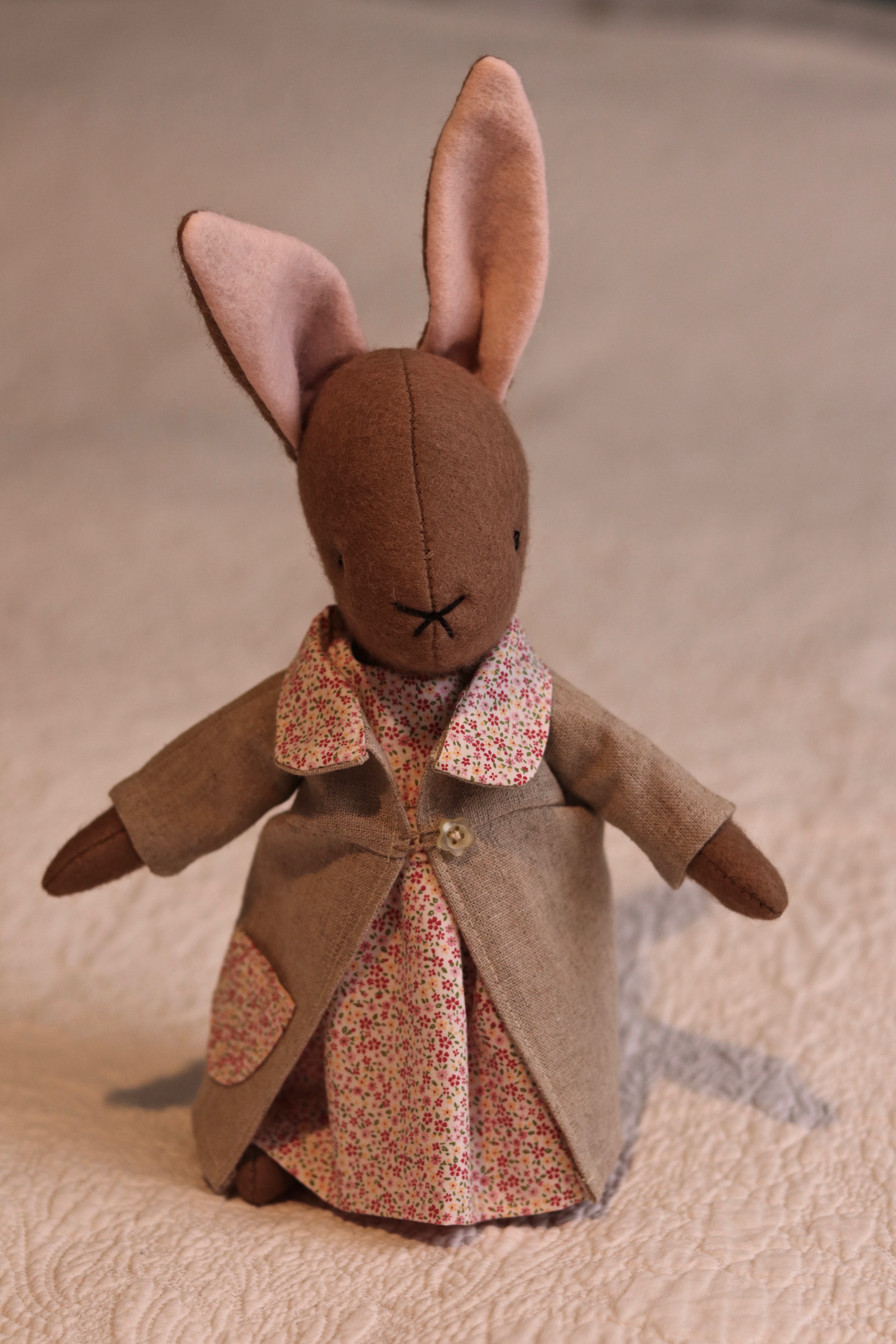 A handmade brown felt bunny rabbit with cotton floral dress and a natural linen coat with a hand embroidered name on her ear. Made using a pattern by Simone Gooding.