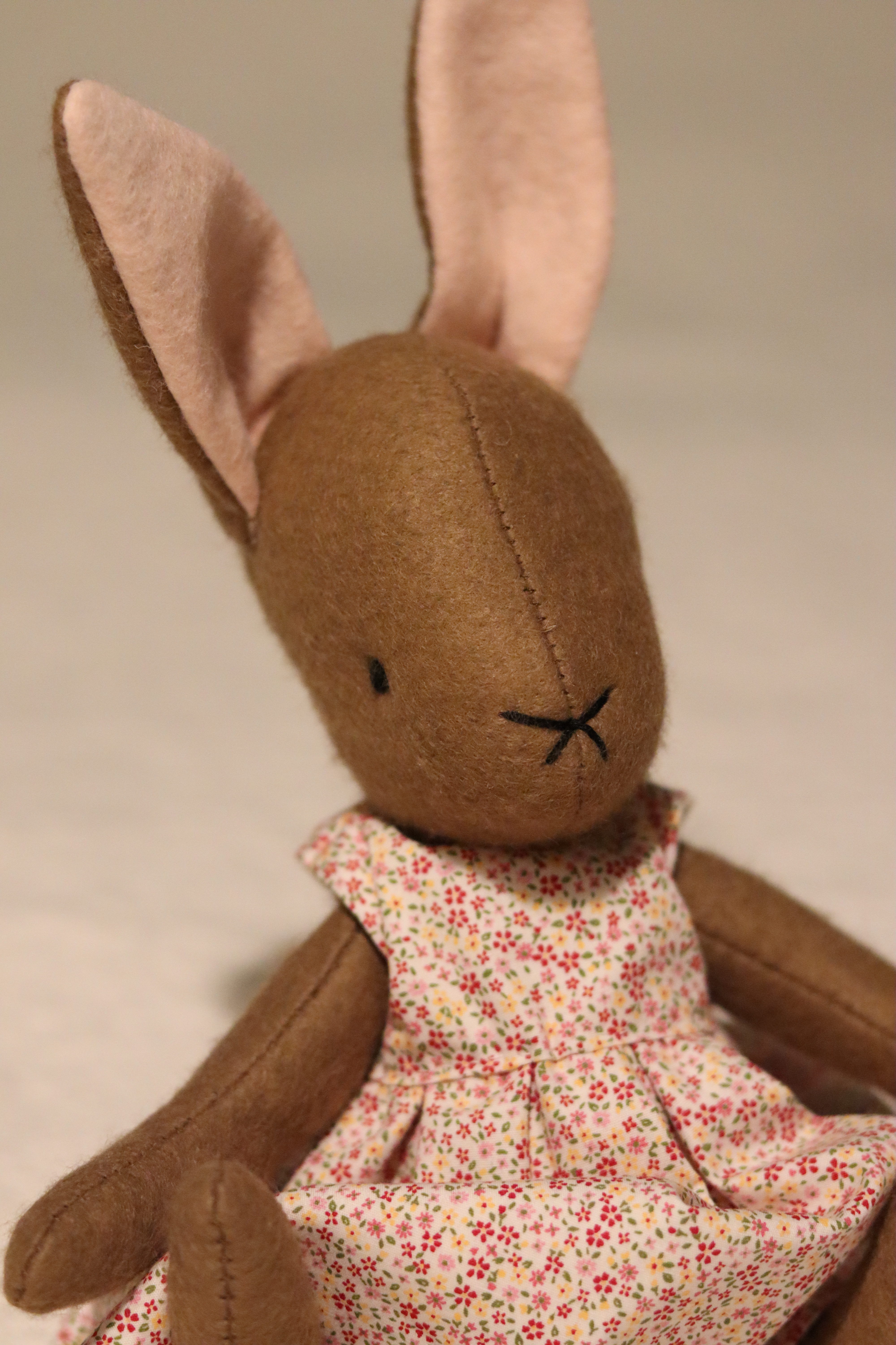 A handmade brown felt bunny rabbit with cotton floral dress and a natural linen coat with a hand embroidered name on her ear. Made using a pattern by Simone Gooding.