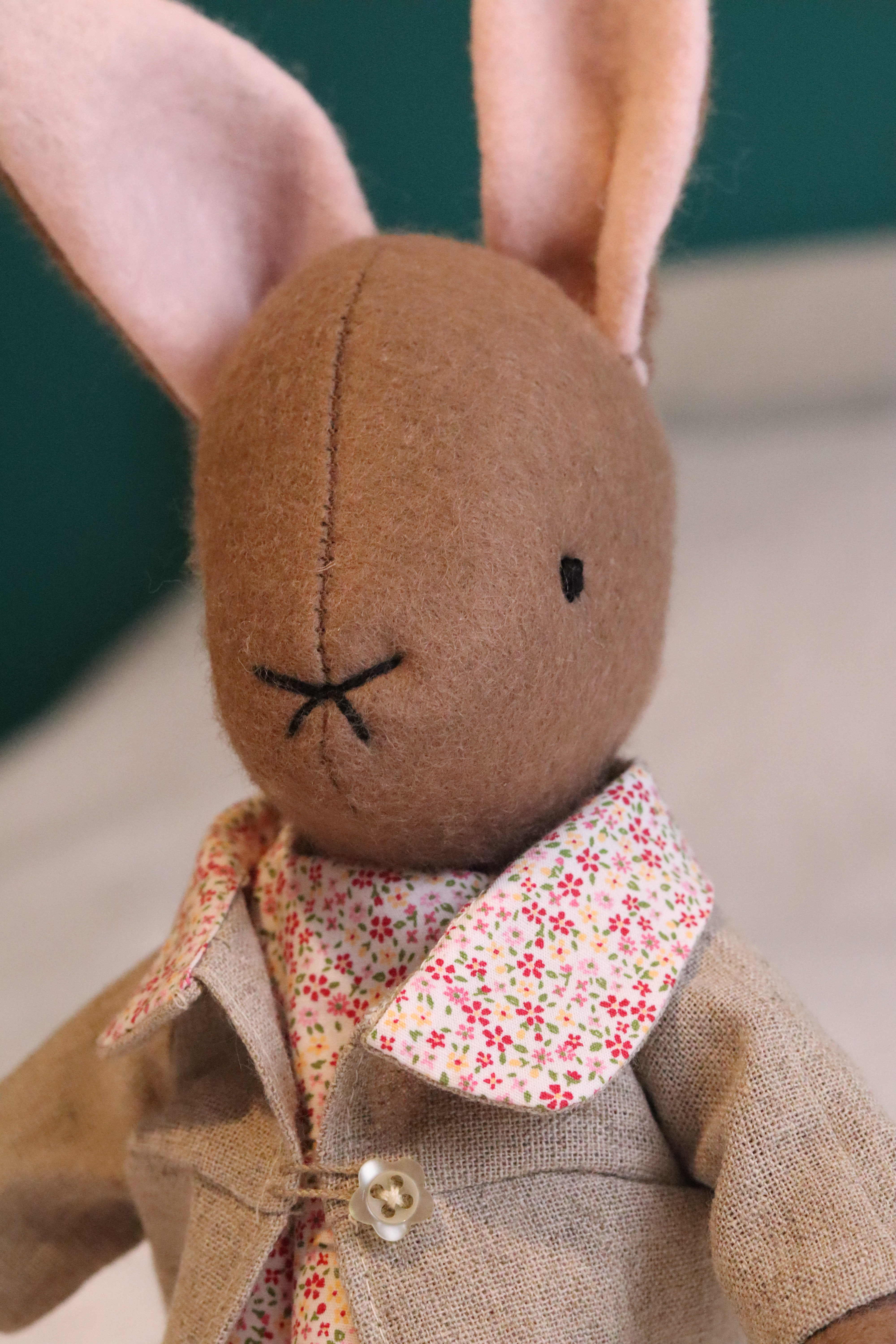 A handmade brown felt bunny rabbit with cotton floral dress and a natural linen coat with a hand embroidered name on her ear. Made using a pattern by Simone Gooding.