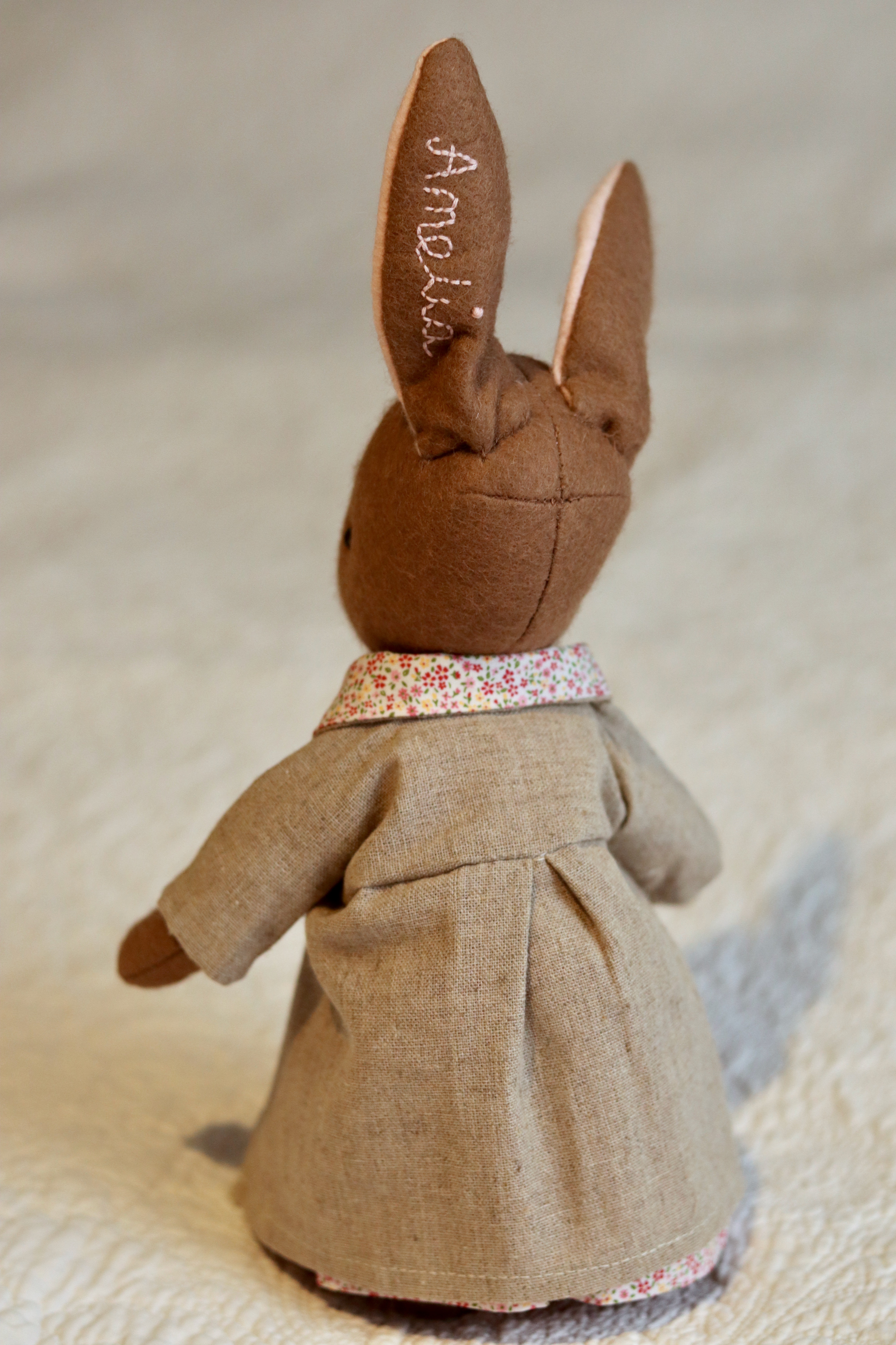 A handmade brown felt bunny rabbit with cotton floral dress and a natural linen coat with a hand embroidered name on her ear. Made using a pattern by Simone Gooding.