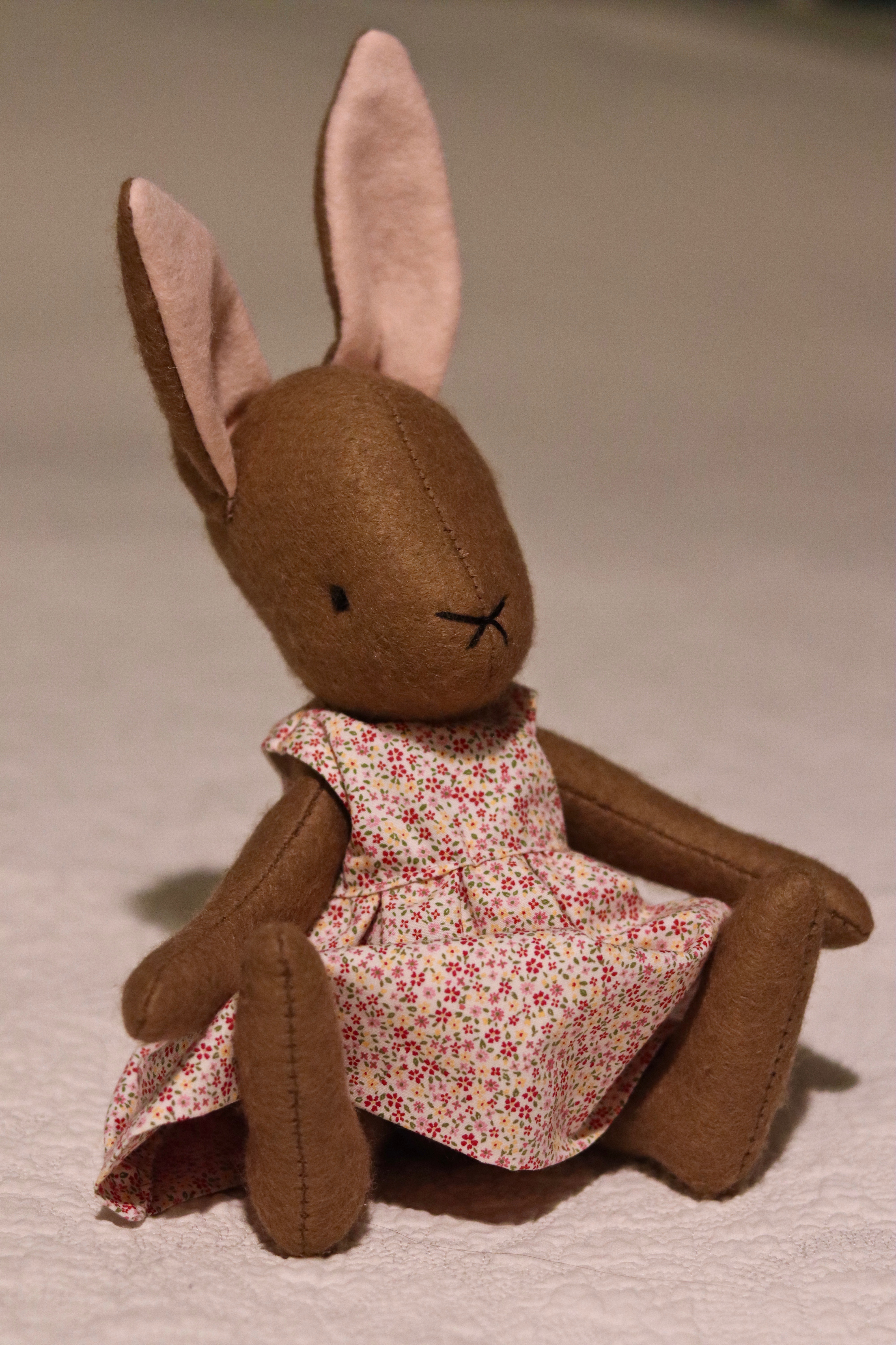 A handmade brown felt bunny rabbit with cotton floral dress and a natural linen coat with a hand embroidered name on her ear. Made using a pattern by Simone Gooding.