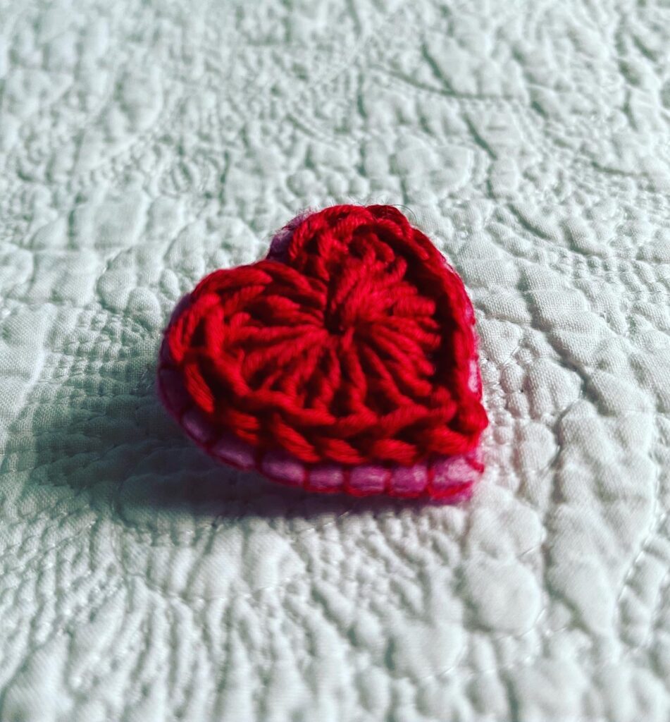 A small red crocheted heart on a pink hand stitched felt back with metal fixing brooch. Approximate size 4cm width x 4cm height.