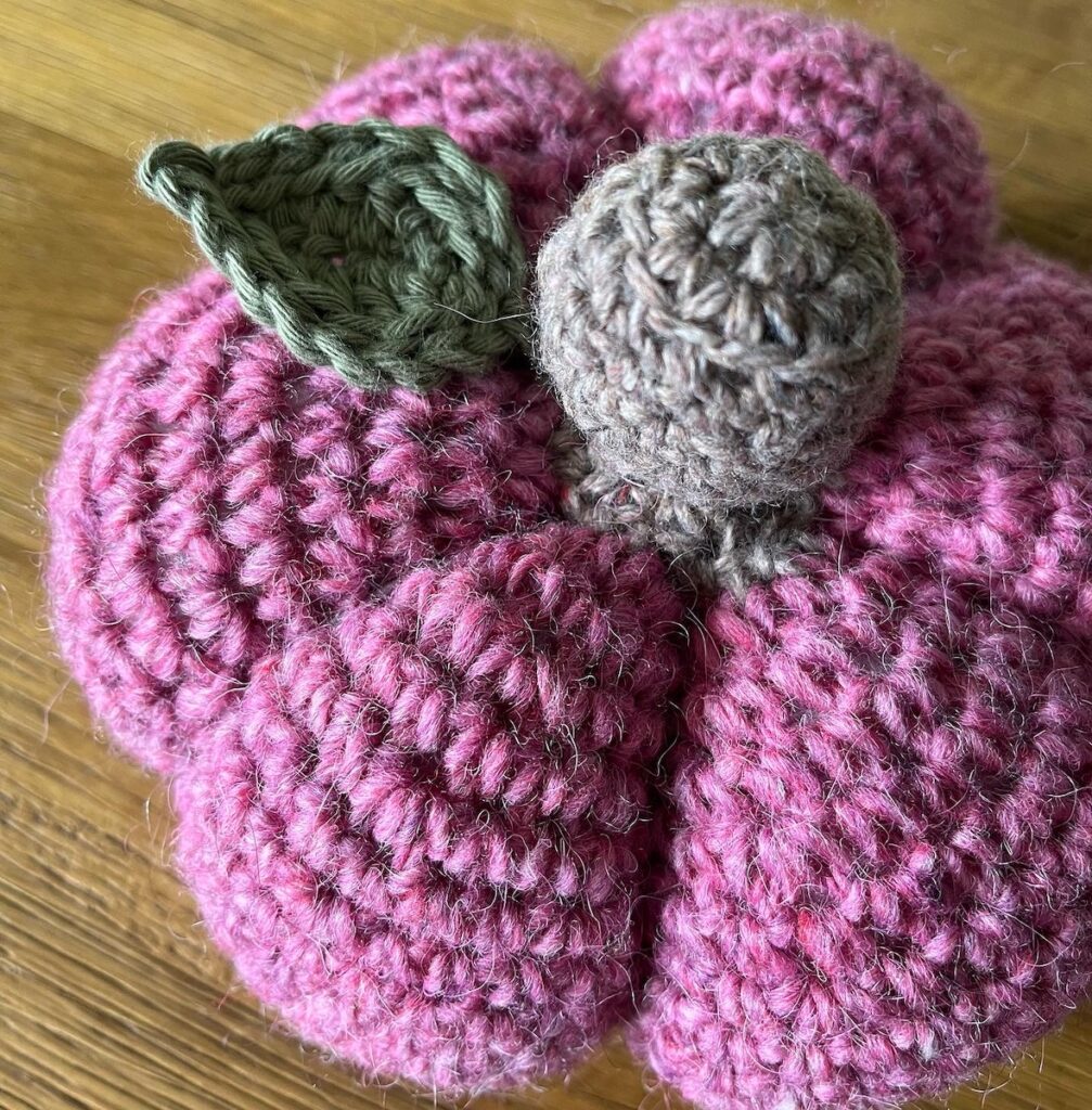 A hand crocheted pumpkin decoration, made using pink pure wool yarn with a brown wool stem and green cotton leaf detail.