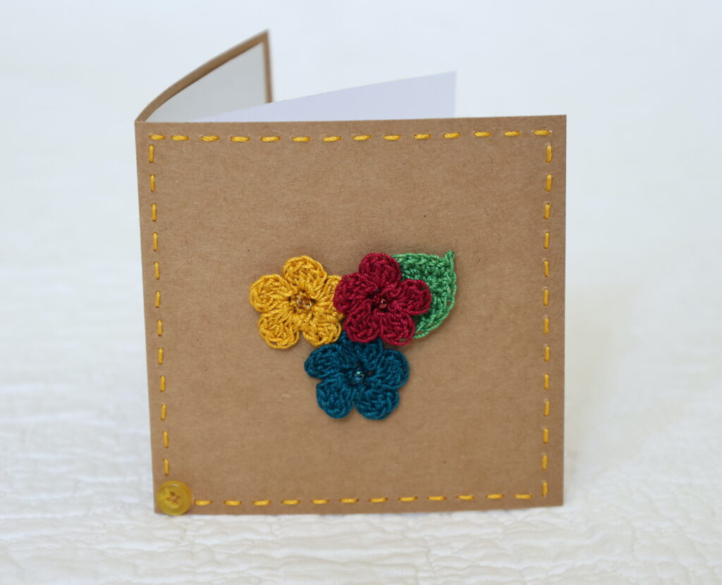 Small brown square greetings card with three crocheted flowers and leaf in centre and hand stitched border. 