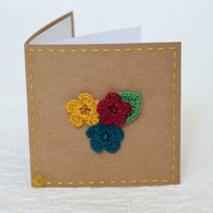 Small brown square greetings card with three crocheted flowers and leaf in centre and hand stitched border.
