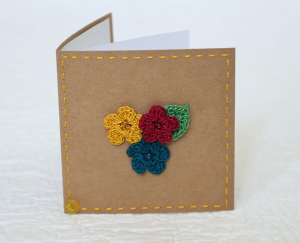 Small brown square greetings card with three crocheted flowers and leaf in centre and hand stitched border.