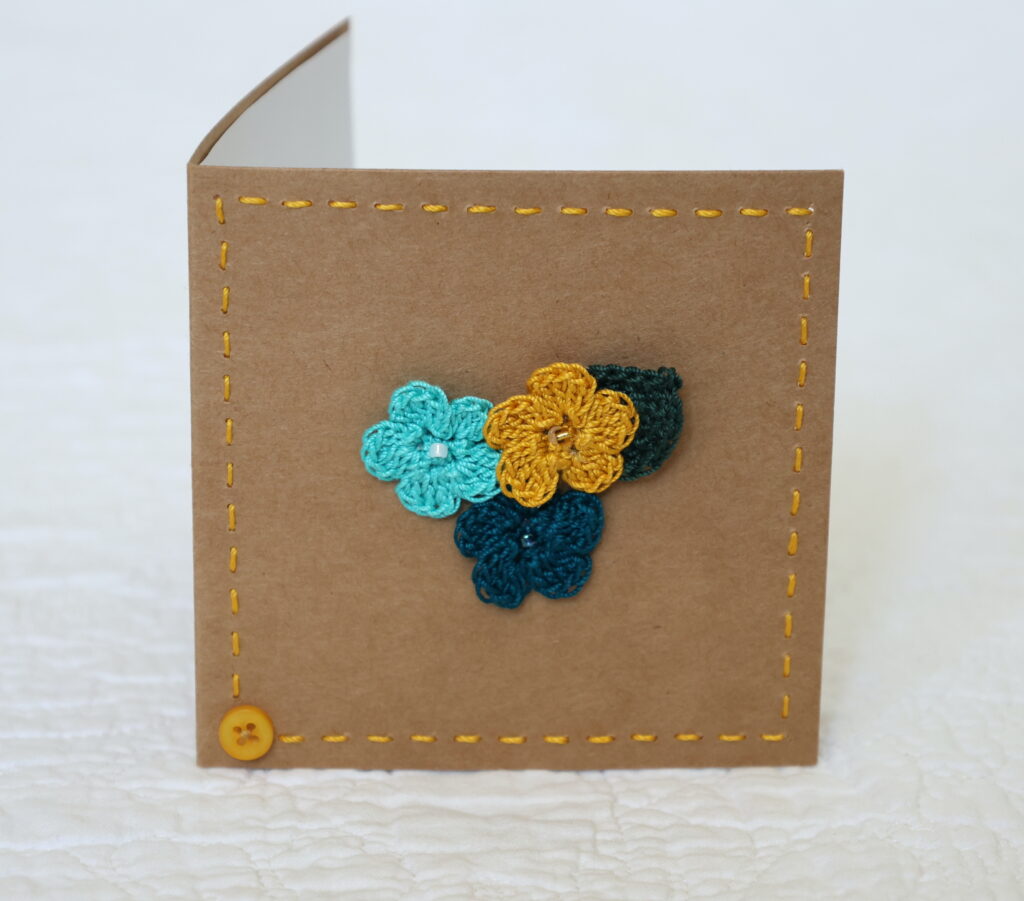 Posy of 3 crocheted flowers and a leaf with hand stitched border on a small brown card (approximate size 10 x 10 cm)with blank white insert. Brown envelope included.