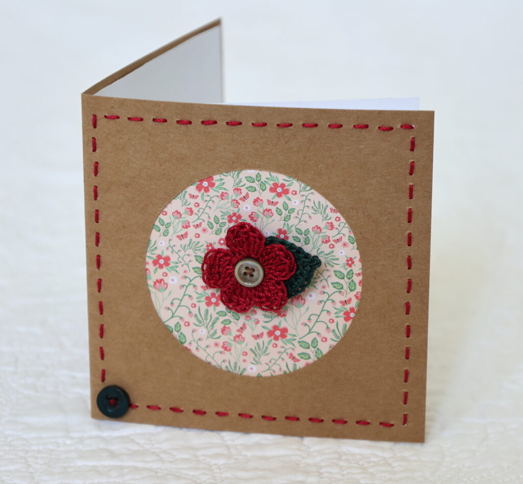 A single crocheted flower and a leaf with hand stitched border on a small brown card (approximate size 10 x 10 cm)with blank white insert. Brown envelope included.