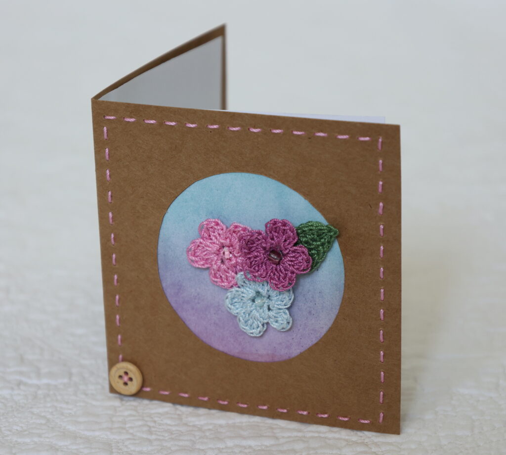 Posy of 3 crocheted flowers and a leaf with hand stitched border on a small brown card (approximate size 10 x 10 cm)with blank white insert. Brown envelope included.