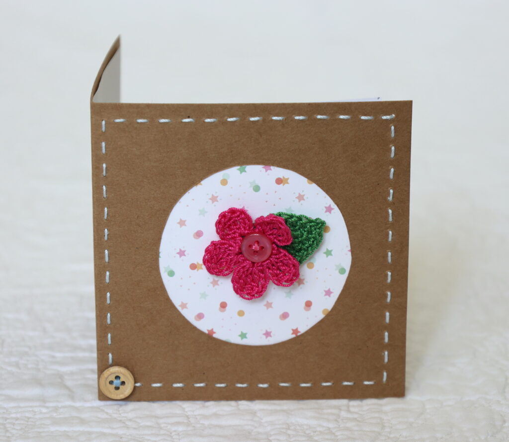 A single crocheted flower and a leaf with hand stitched border on a small brown card (approximate size 10 x 10 cm)with blank white insert. Brown envelope included.