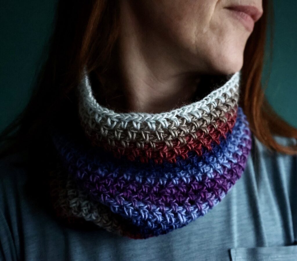 A tube style neck warmer in a mixture of muted pink, purple, and blue shades. Made using a fine, soft and lightweight acrylic/wool mix yarn.