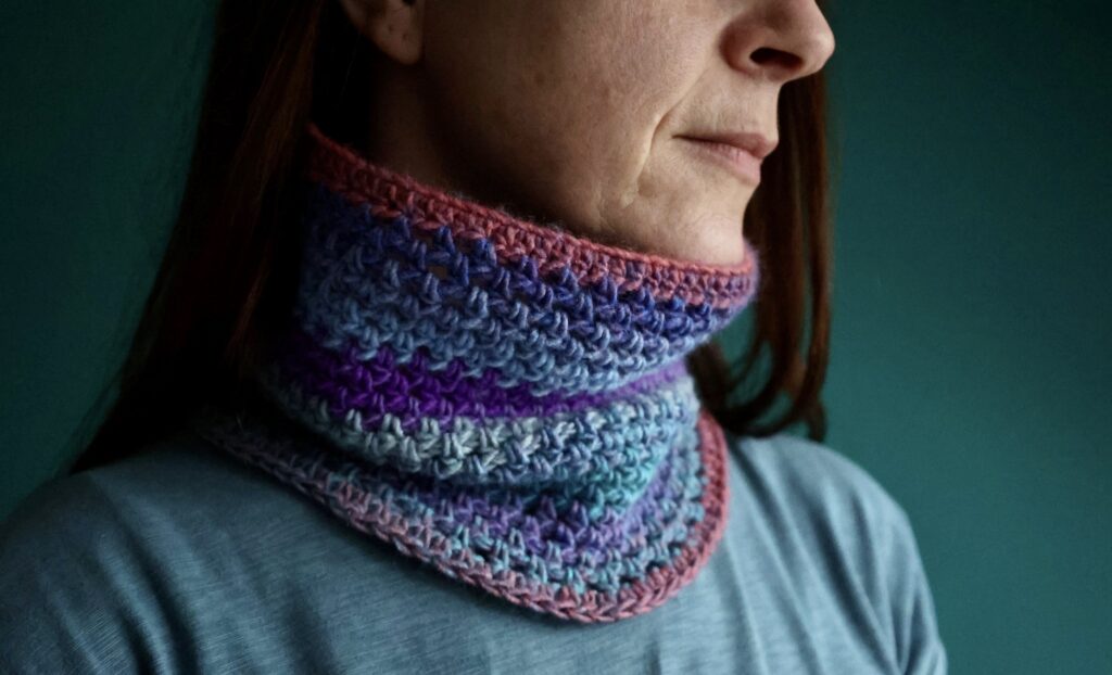 A tube style neck warmer in a mixture of muted pink, purple, and blue shades. Made using a fine, soft and lightweight acrylic/wool mix yarn.