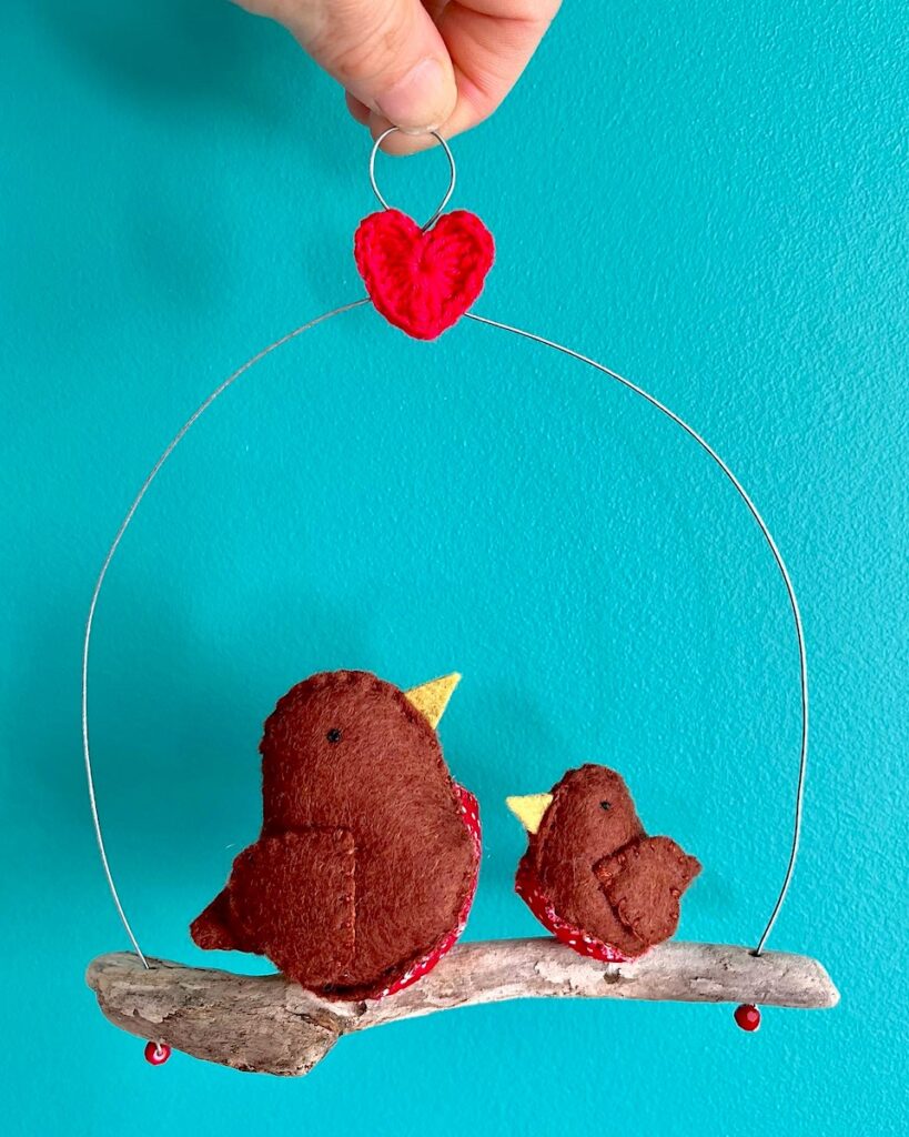 A pair of little birds (a single small and single medium sized), handmade in brown felt with a red cotton spotty print fabric chest. These birds are sat on a natural driftwood perch with a wire hanger that is decorated with a crocheted red heart.