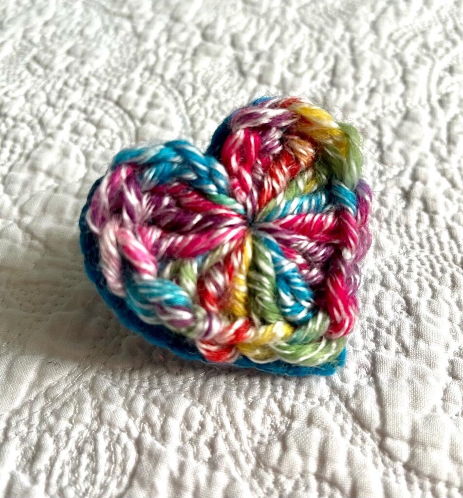 A bright, multicoloured crocheted heart on a turquoise hand stitched felt back with metal fixing brooch. Approximate size 5.5cm width x 5.5cm height.