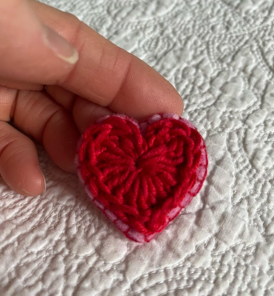 A small red crocheted heart on a pink hand stitched felt back with metal fixing brooch. Approximate size 4cm width x 4cm height.