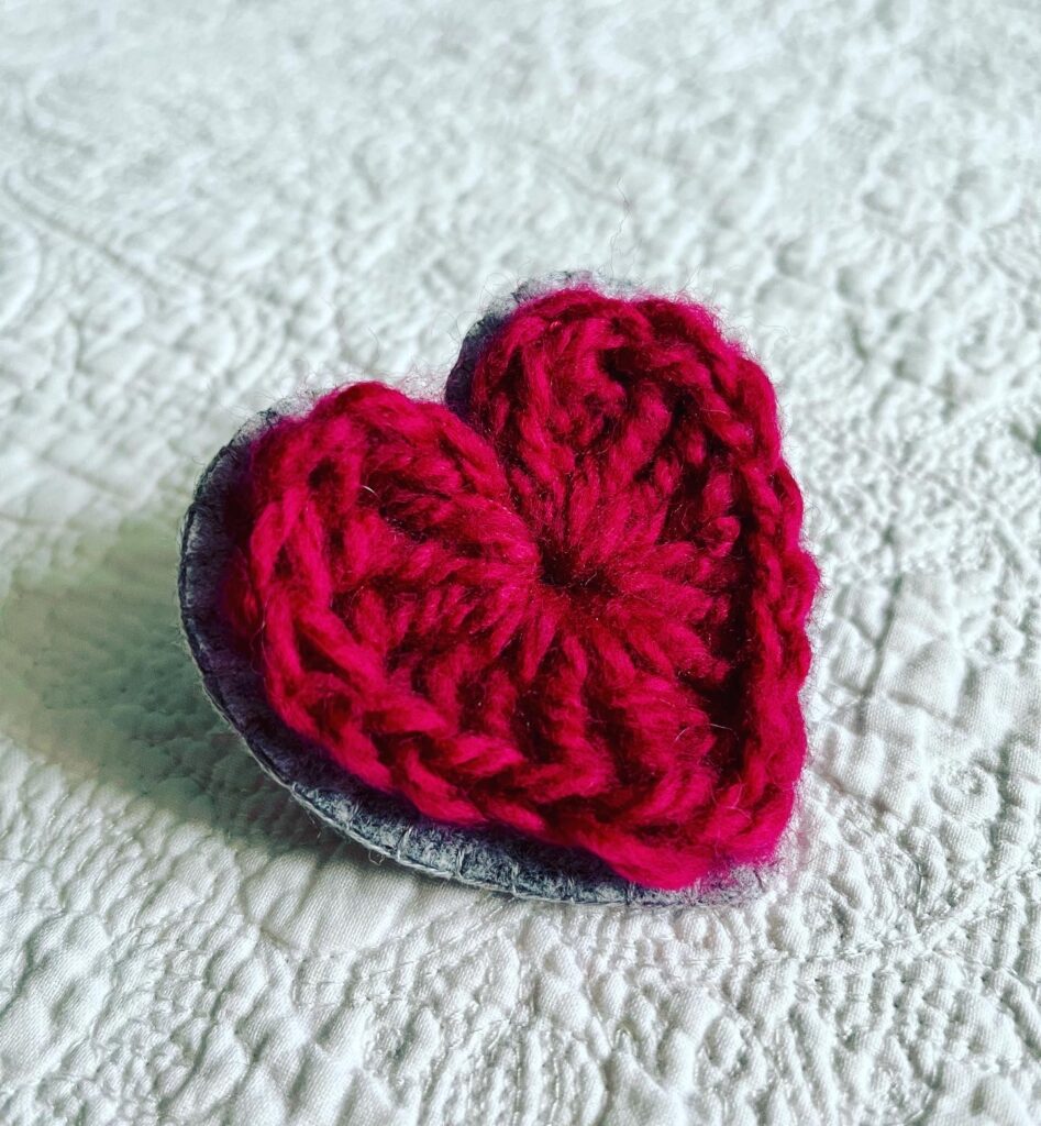 A crocheted red heart on a grey hand stitched felt back with metal fixing brooch. Approximate size 7cm width x 7cm height.