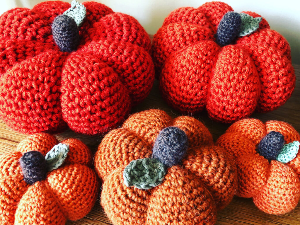 Crochet pumpkins in a selection of sizes, thickness and texture of yarn or wool. In warm tones of orange with a brown stalk and green leaf detail.