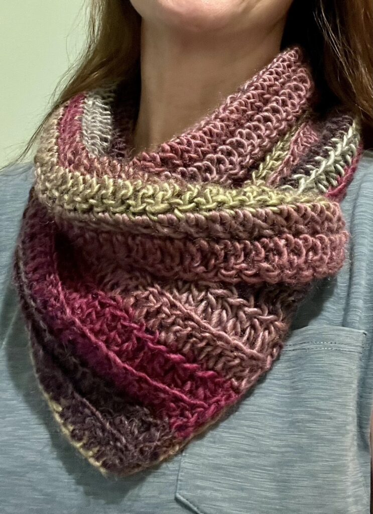 A cross over style neck warmer in a mixture of muted pink, purple, and green hues. Made using a chunky, soft and lightweight acrylic/wool mix yarn.