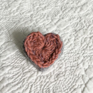 A small pink crocheted heart on a grey hand stitched felt back with metal fixing brooch. Approximate size 4cm width x 4cm height.