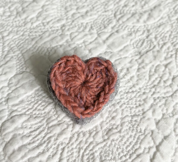A small pink crocheted heart on a grey hand stitched felt back with metal fixing brooch. Approximate size 4cm width x 4cm height.