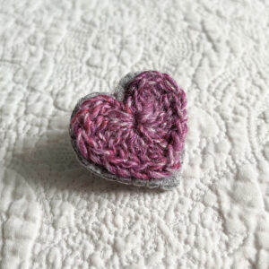 A small purple crocheted heart on a grey hand stitched felt back with metal fixing brooch. Approximate size 4cm width x 4cm height.