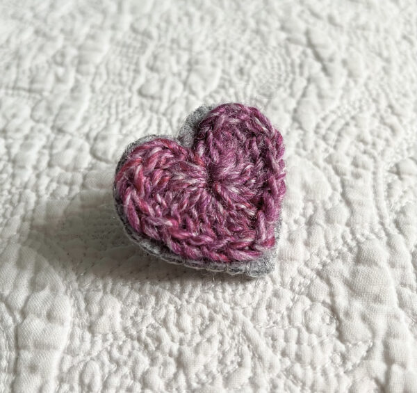 A small purple crocheted heart on a grey hand stitched felt back with metal fixing brooch. Approximate size 4cm width x 4cm height.
