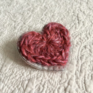 A crocheted red heart on a grey hand stitched felt back with metal fixing brooch. Approximate size 6.5cm width x 6.5cm height.