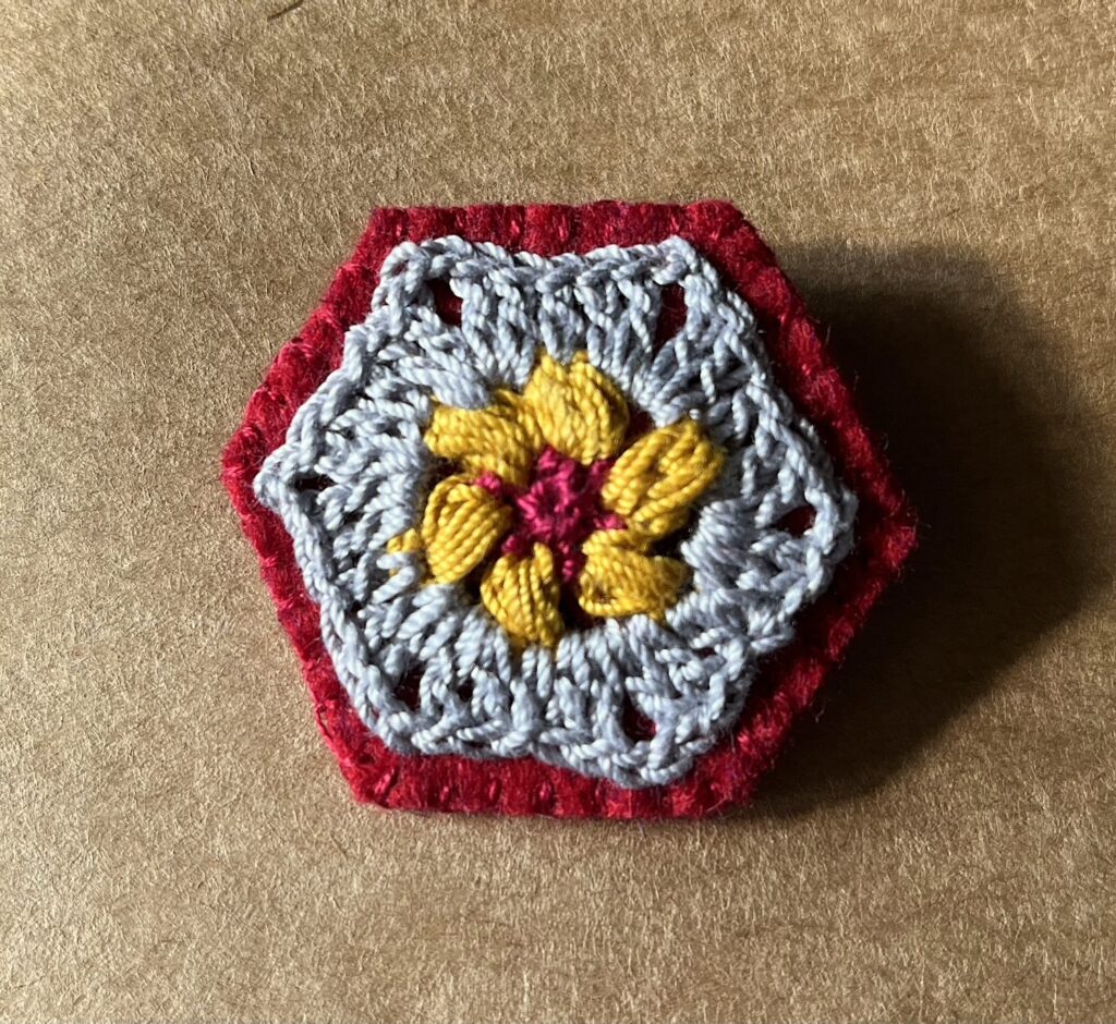 A hexagon shaped, hand stitched, felt and flower design crocheted brooch with metal locking brooch fixing.